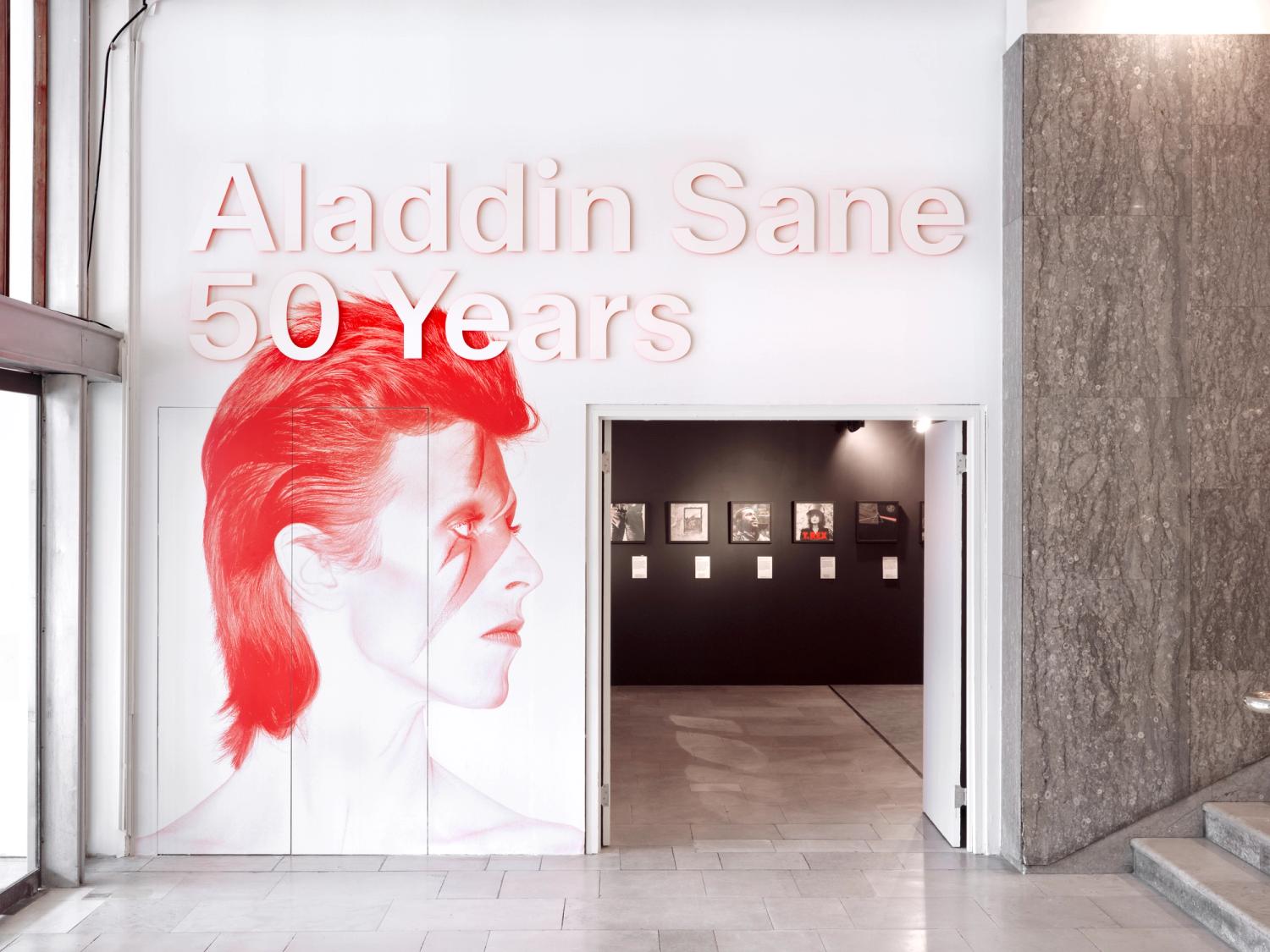 A large wall with a wide doorway leading to a dark gallery. On the wall is a duotone image of David Bowie in red, and the exhibition title.