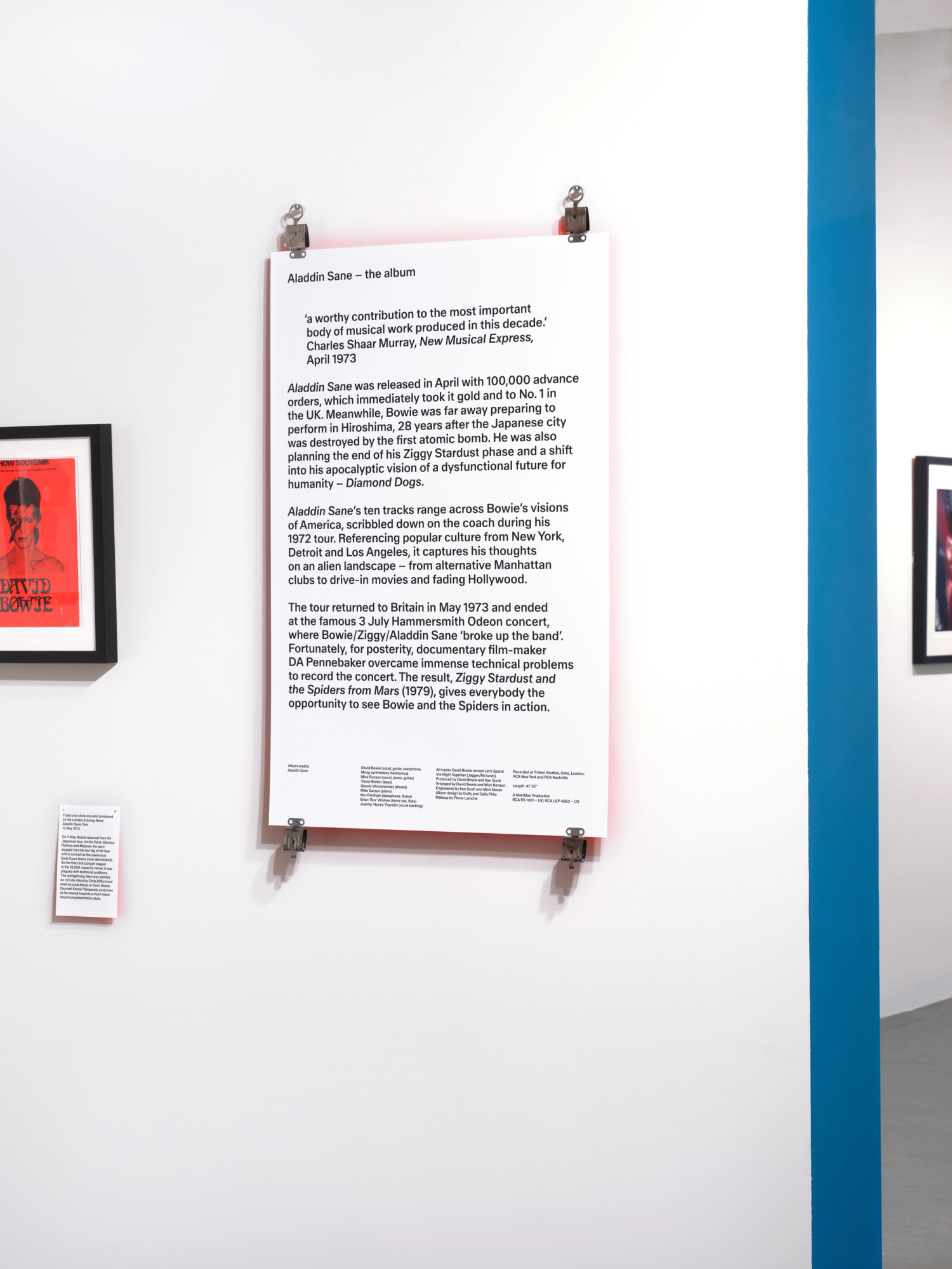 A text panel in a gallery space. The panel has a red glow reflecting against the white wall. It is secured with stainless steel clips at each corner.