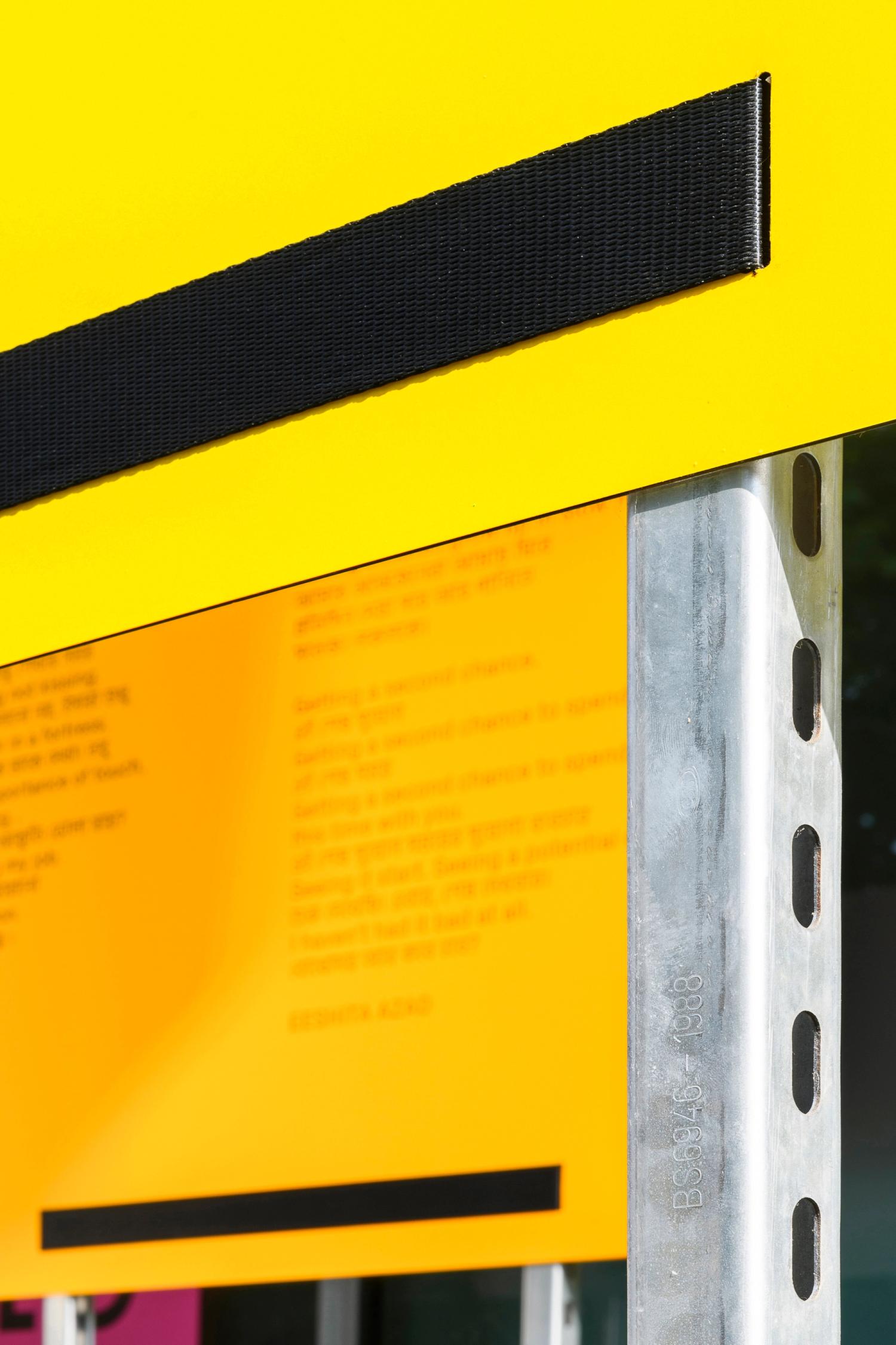 A close-up detail of a yellow panel fixed using black seatbelt strapping to a stainless steel vertical bar.