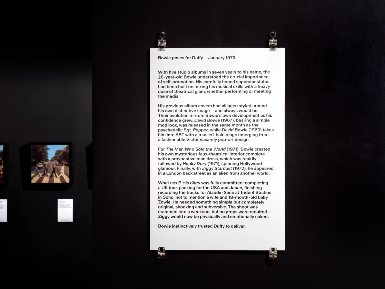 A white text panel pinned to the wall in a gallery painted black. The panel is secured with large stainless steel clips at the four corners. 