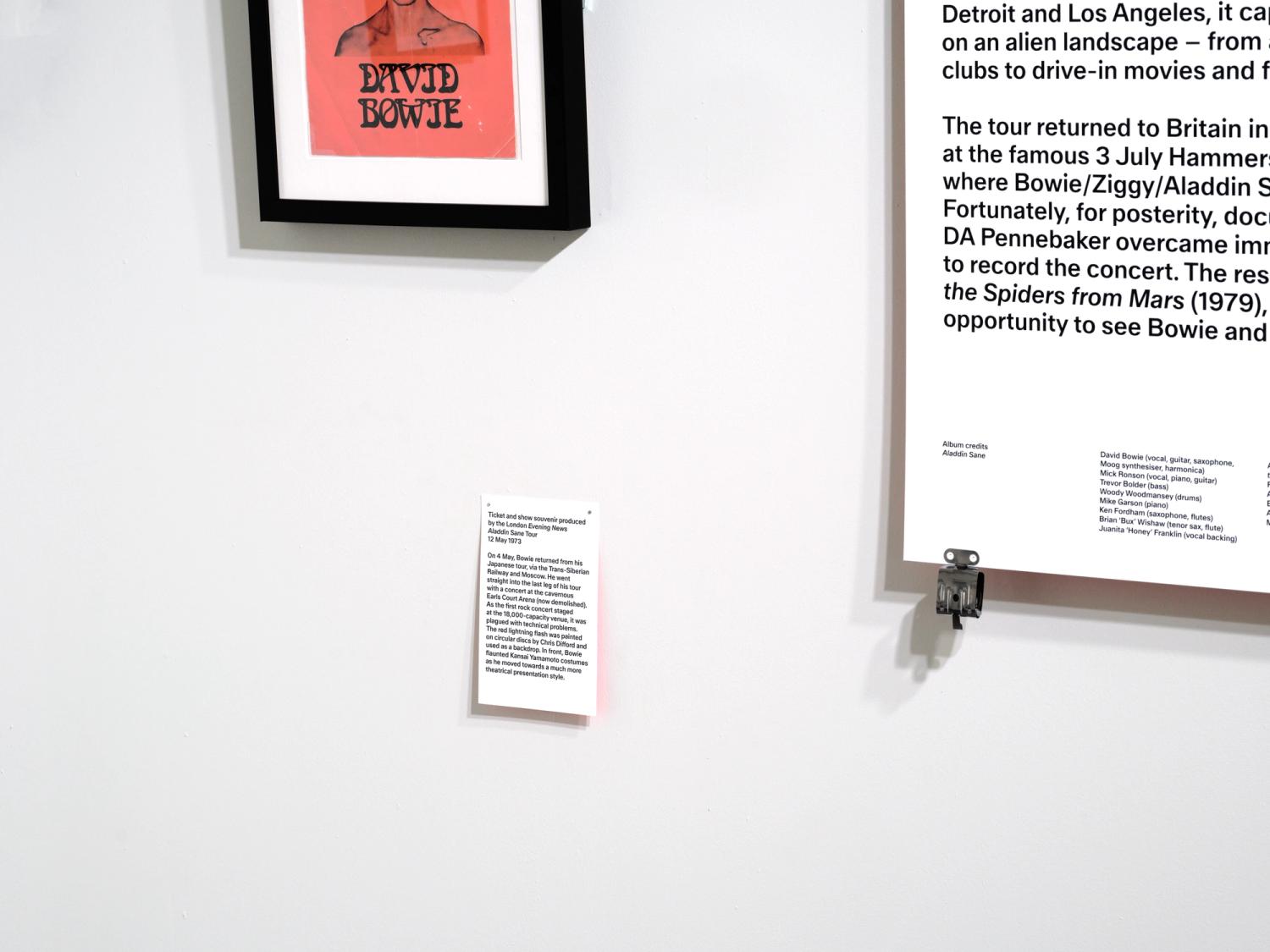 An artwork caption pinned to the wall below a red print that reads 'David Bowie', and a larger text panel with a stainless steel clip.