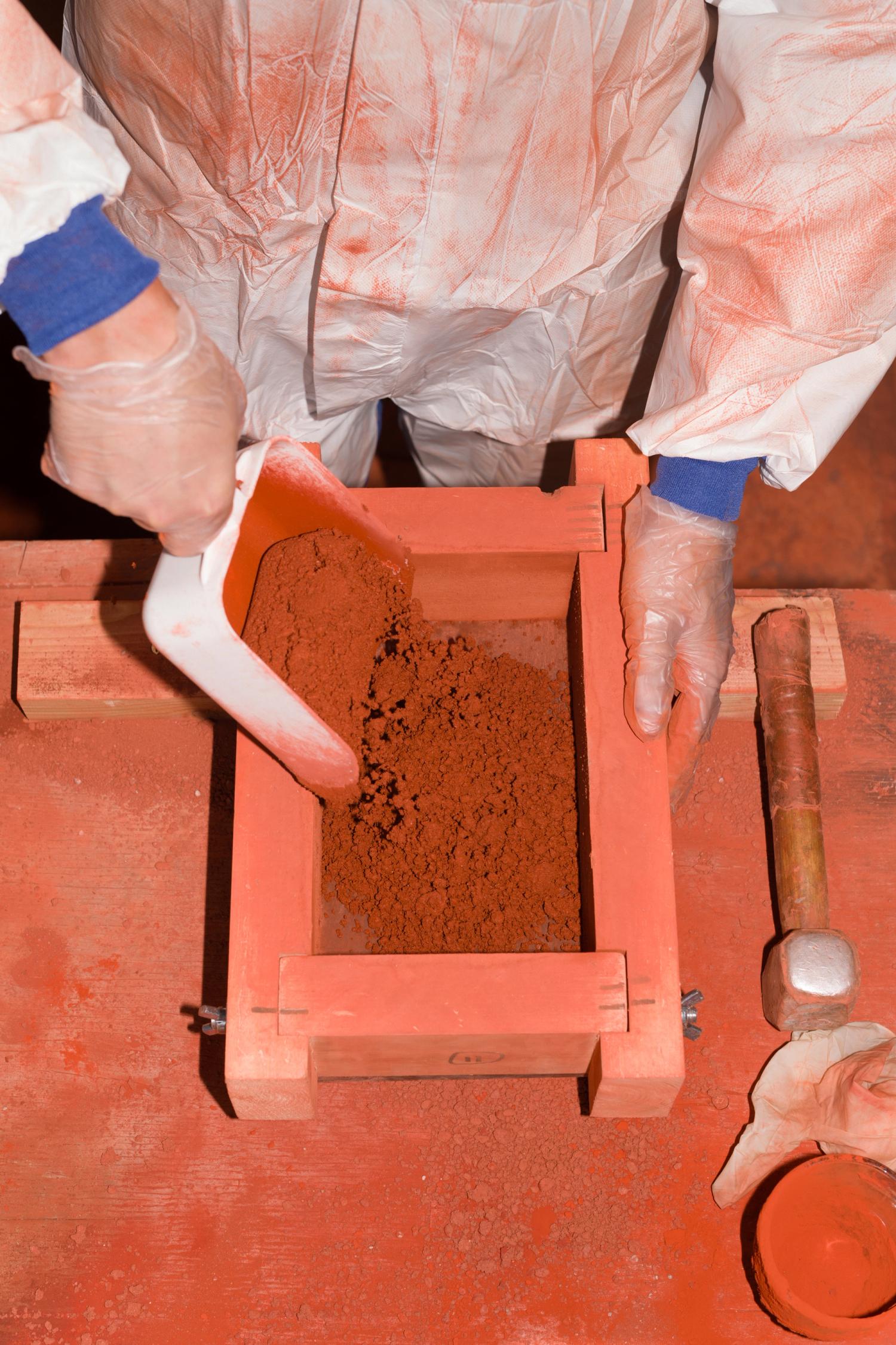 A man in overalls scoops red powder into a wooden rectangular mould