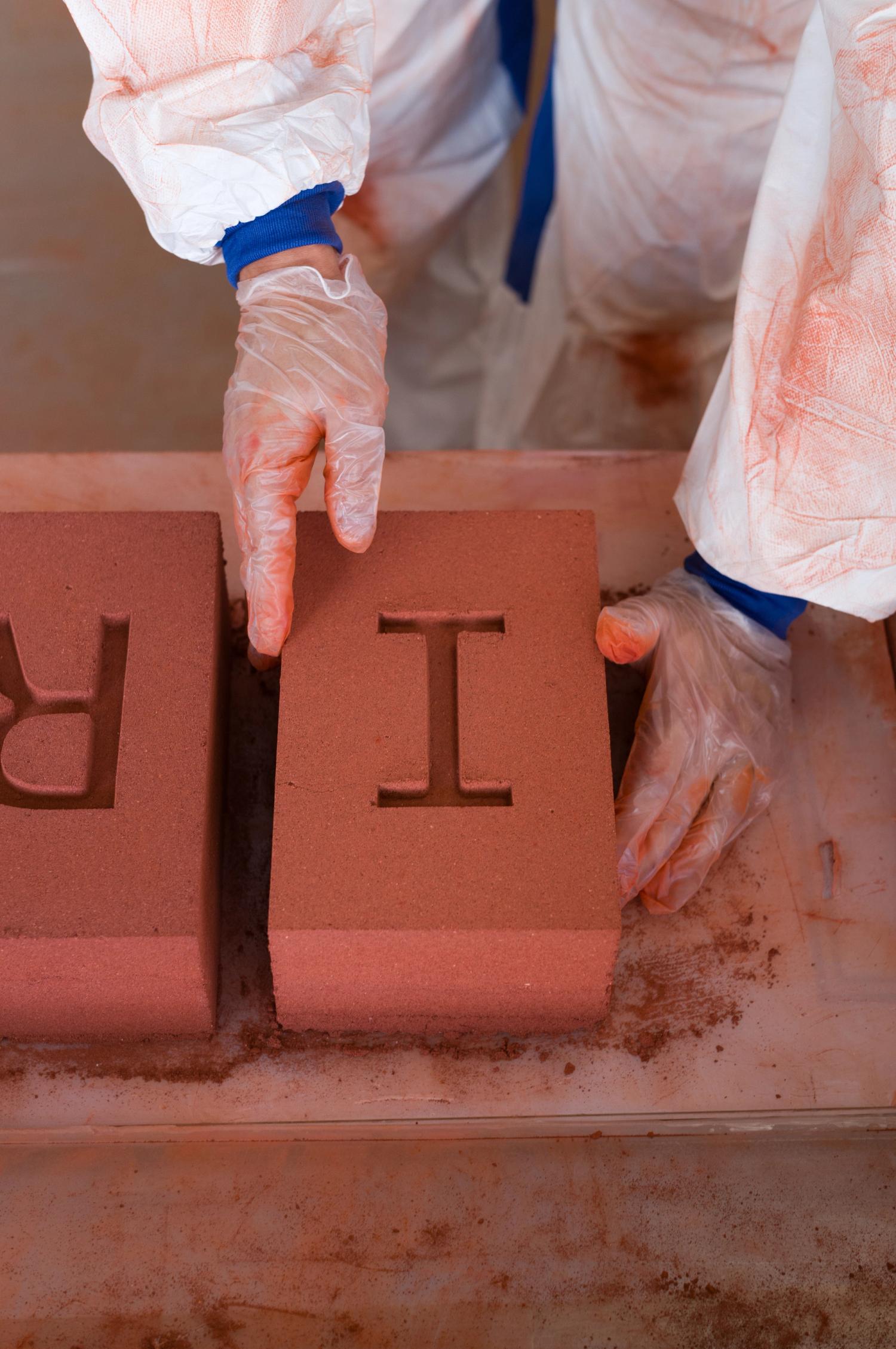 Two gloved hands touching a large red brick with the letter 'I' stamped into it