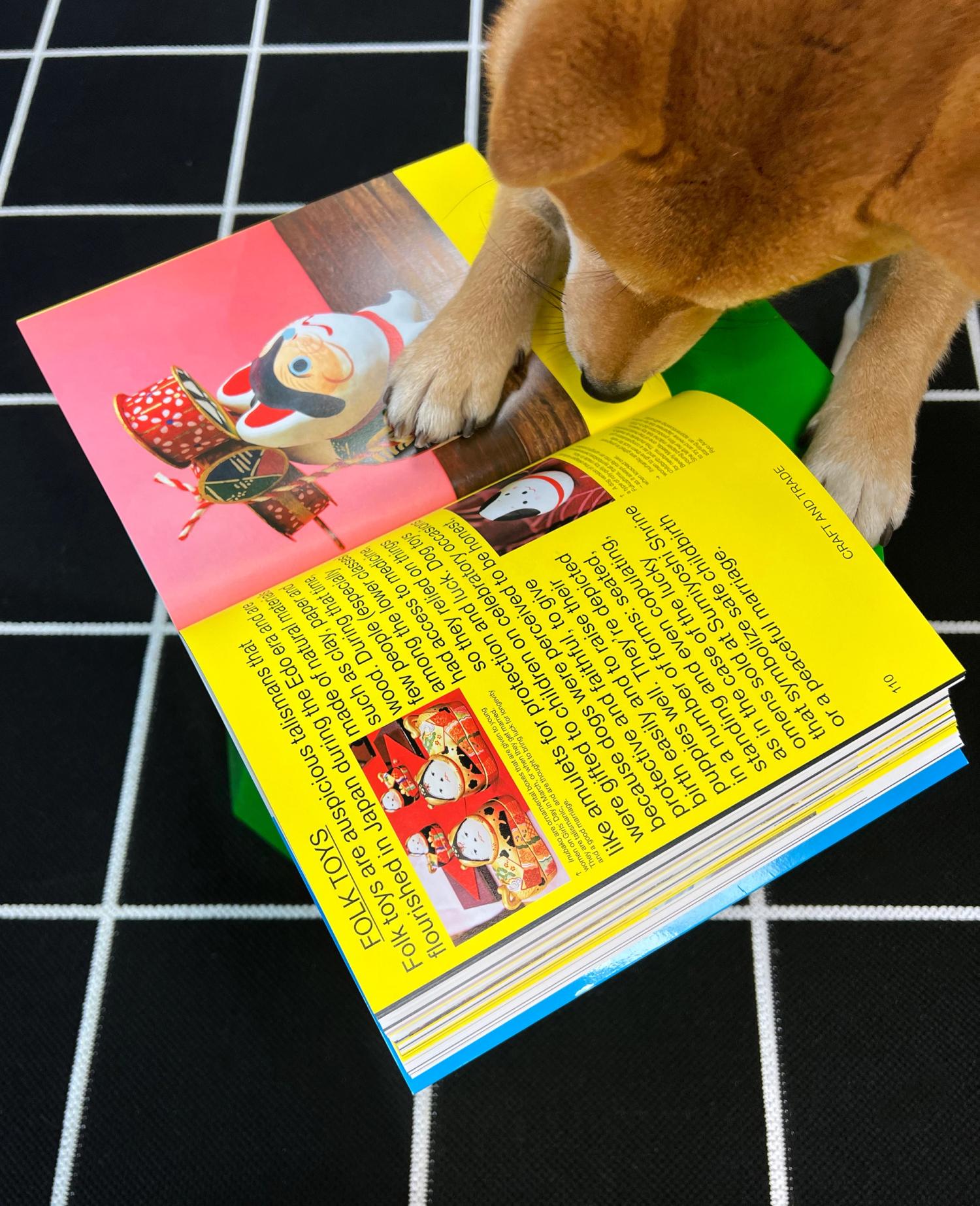 Dog paws on an open book page featuring colorful Japanese dog figurines and text about folk toys, on a black grid background.