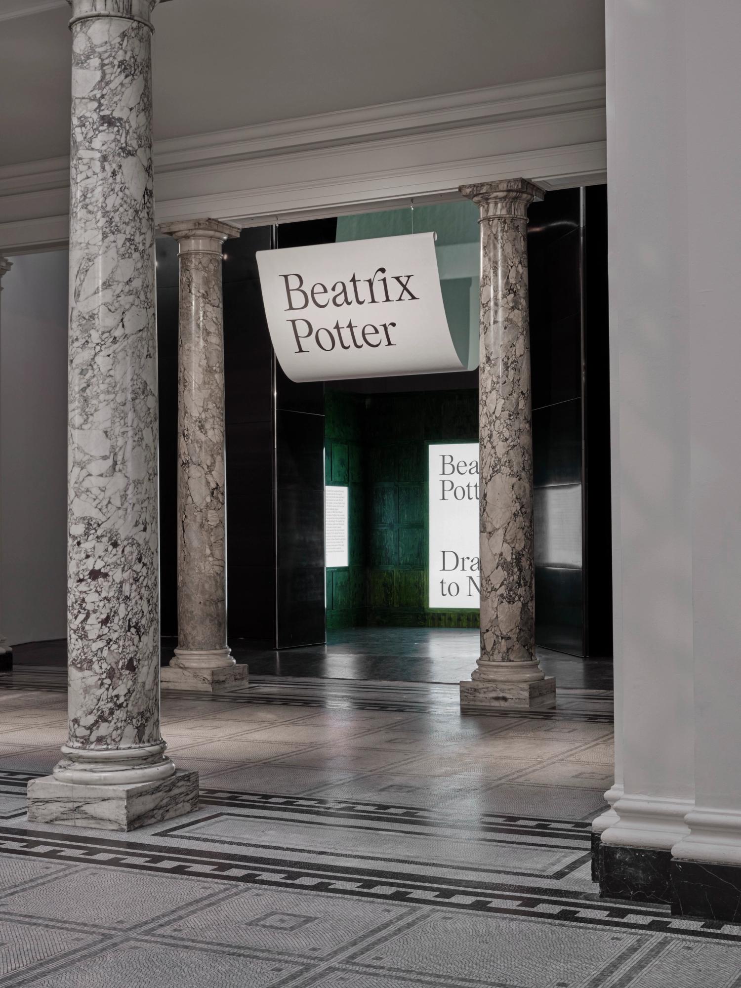 A photograph of a grand entrance to a gallery space within a museum, with marble columns and mosaic floor. A large printed graphic panel hangs in the threshold and a glowing panel is visible within