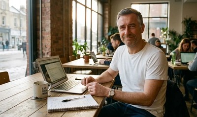 Man in white t-shirt at co-working space table with laptop, calculating mortgage figures in winter sunlight