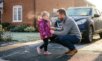 Father in light knit sweater kneels on driveway of modern house, helping daughter with coat in winter sunlight