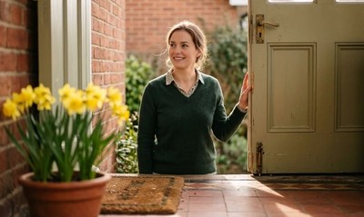 Woman in a jumper stands smiling at the open front door of a bungalow in spring morning light