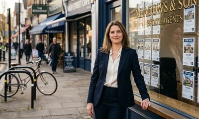 Woman in a suit stands outside an estate agent's window on a high street in spring sunlight
