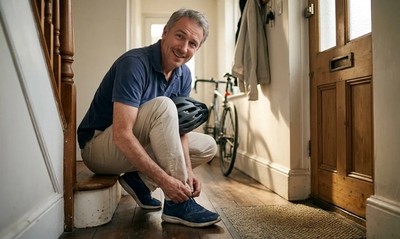 A 57-year-old man ties his trainers in a bright hallway, helmet under his arm, ready for a summer morning cycle.
