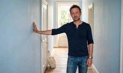 Man in a henley top relaxed in a bright, uncluttered hallway of a converted flat, summer light streaming in