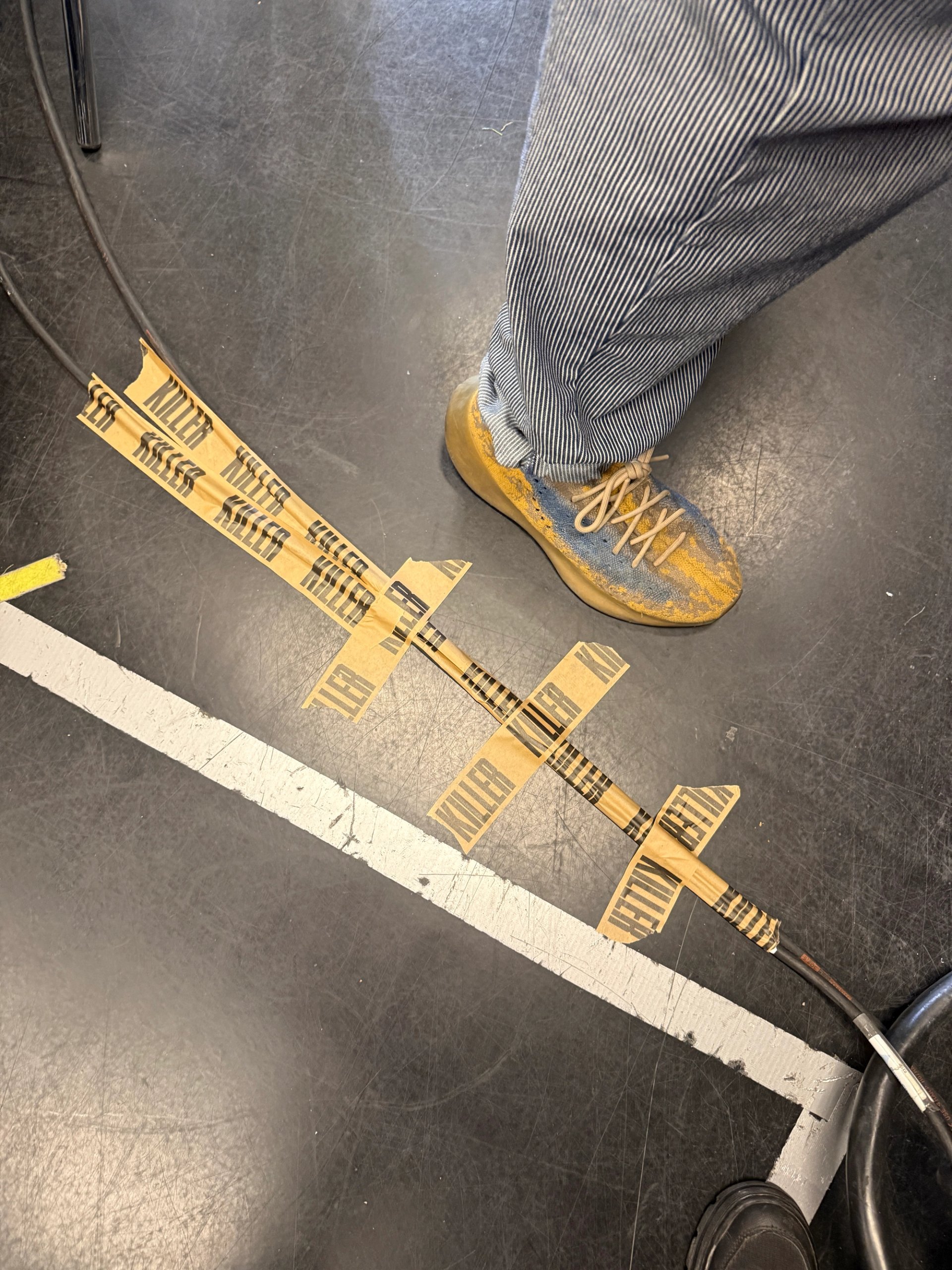 Close-up of KILLER branded gaffer tape marking positions on studio floor next to worn yellow boot