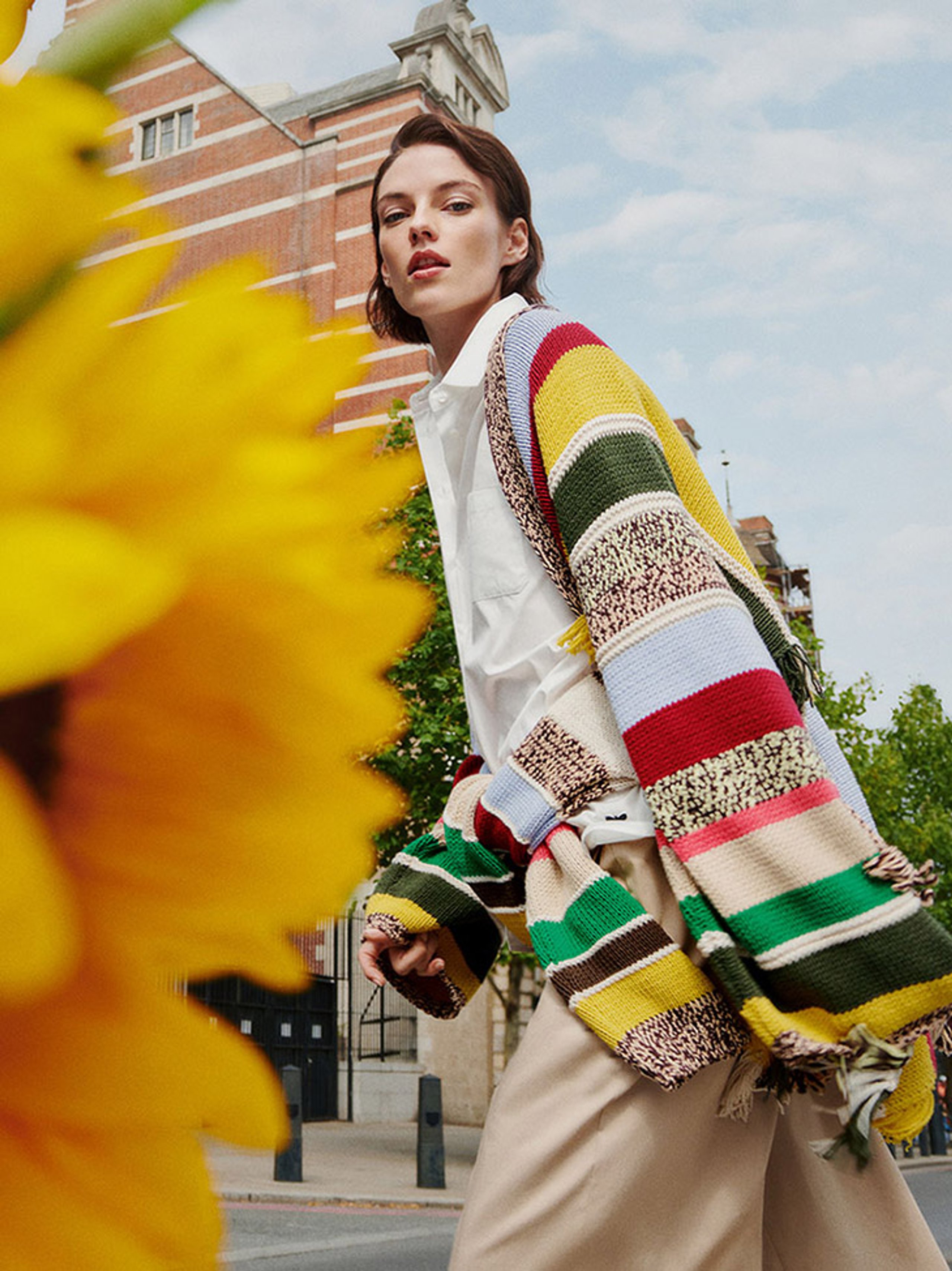 Model in multicolour striped knit cardigan over white shirt with yellow daffodils in foreground