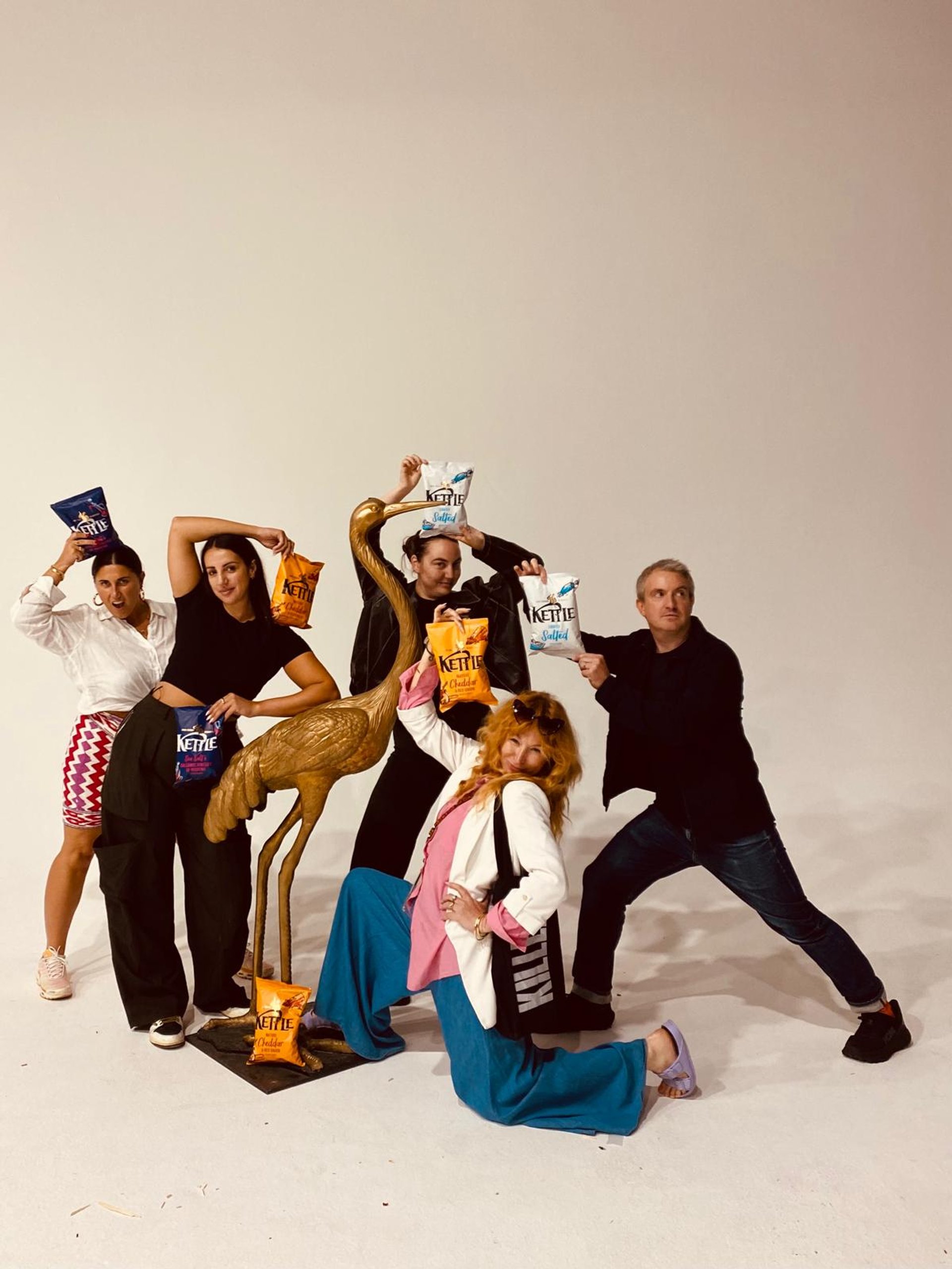 KILLER crew posing with KETTLE Chips packets around gold crane statue on white studio backdrop