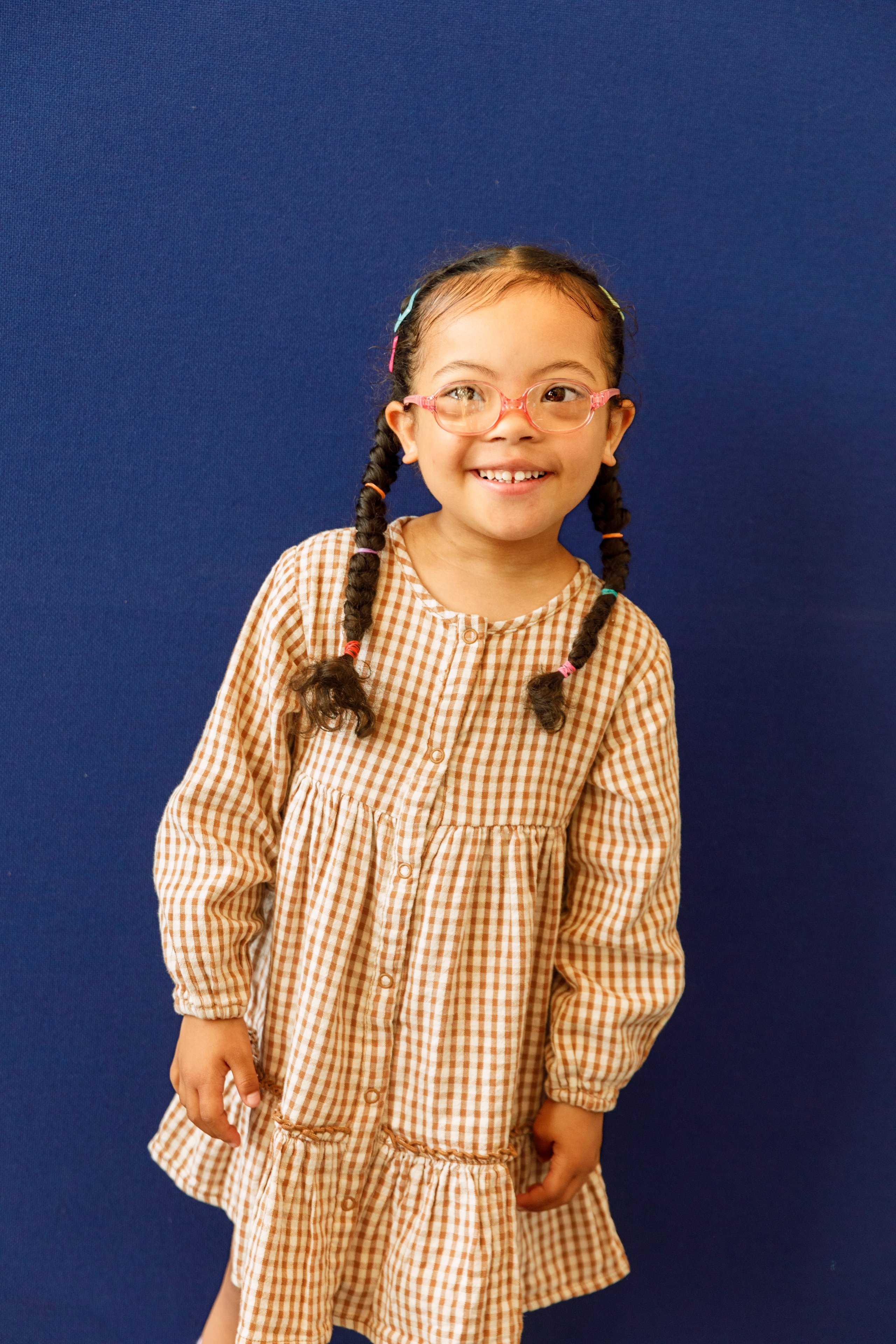 Smiling girl with braids wearing pink glasses and brown gingham tiered dress against navy backdrop