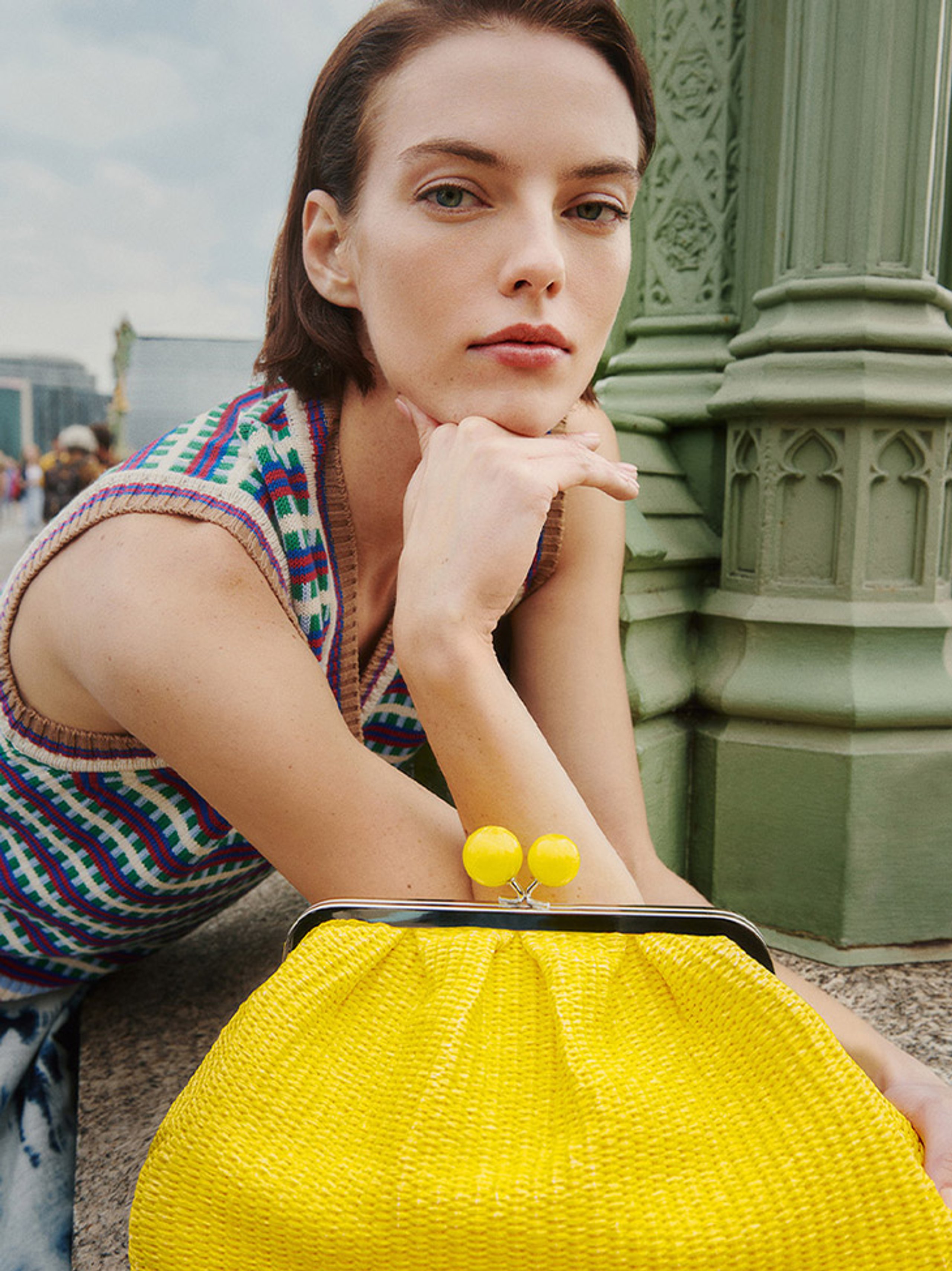 Model leaning on yellow woven clutch bag in multicolour geometric knit vest by ornate green lamppost