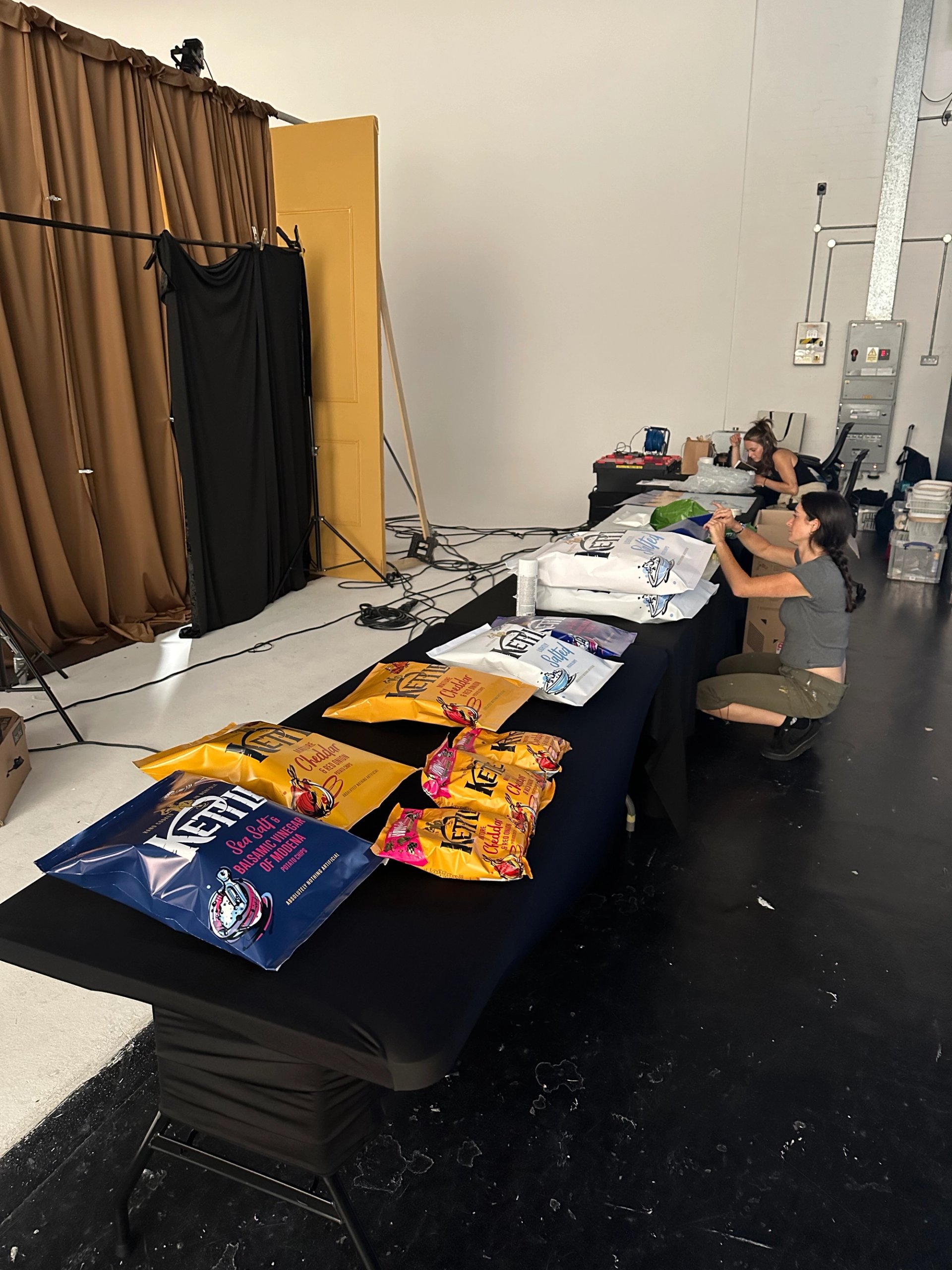 Studio prep table with KETTLE Chips packets laid out and crew preparing wardrobe behind mustard set backdrop