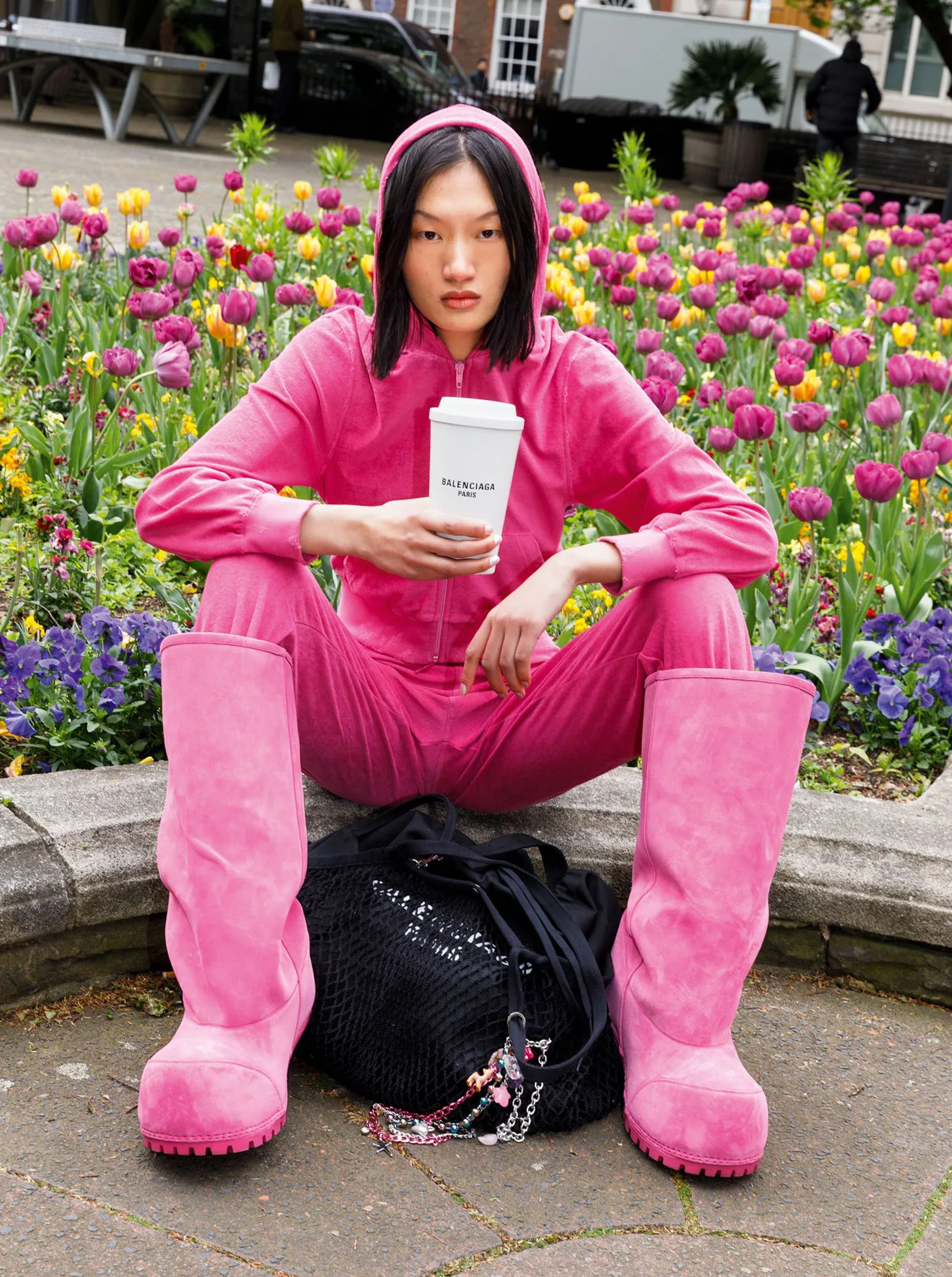 Model in all-pink Balenciaga tracksuit and boots with coffee cup seated by tulip beds