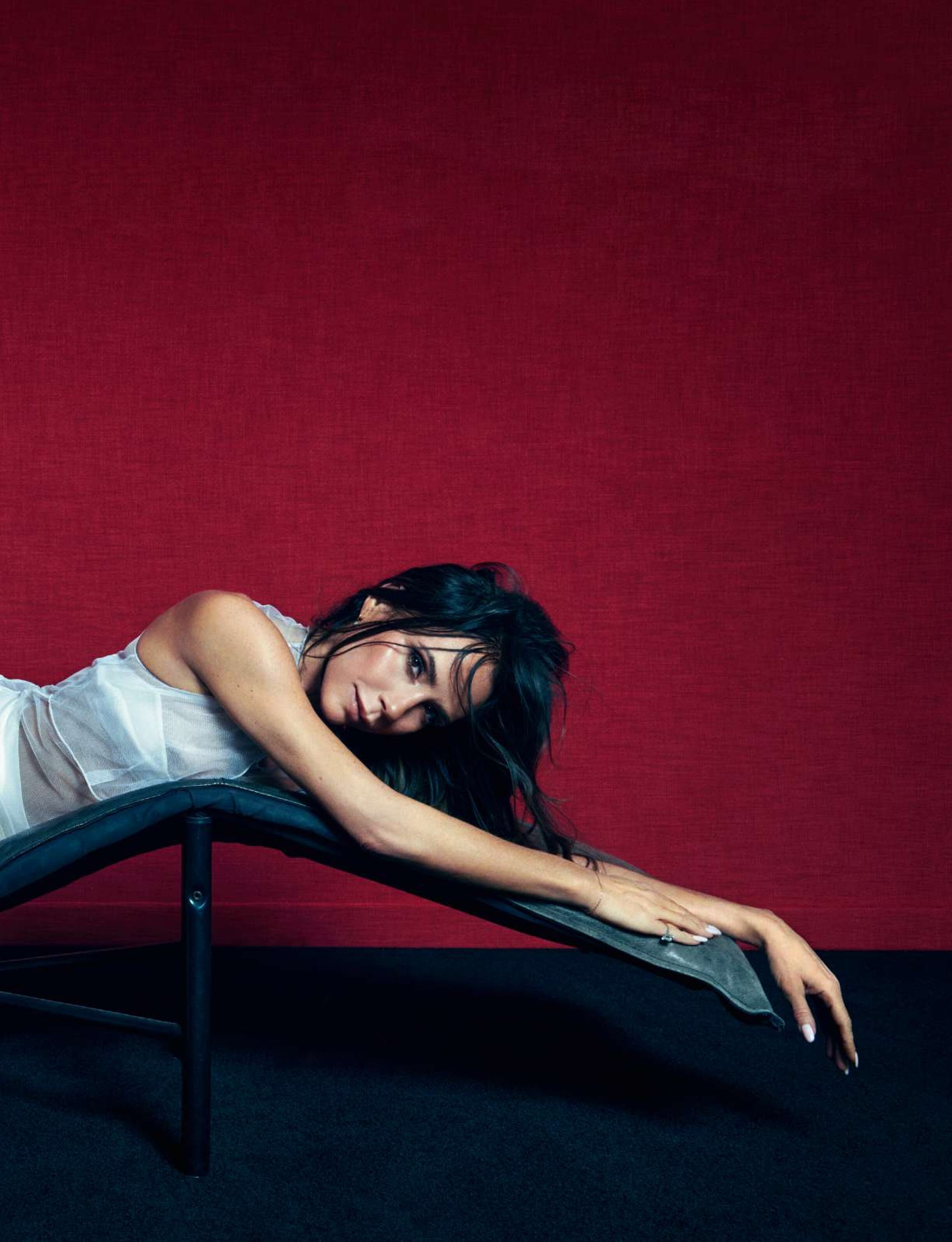 Victoria Beckham lying across leather chaise with tousled hair and white sheer dress against red wall