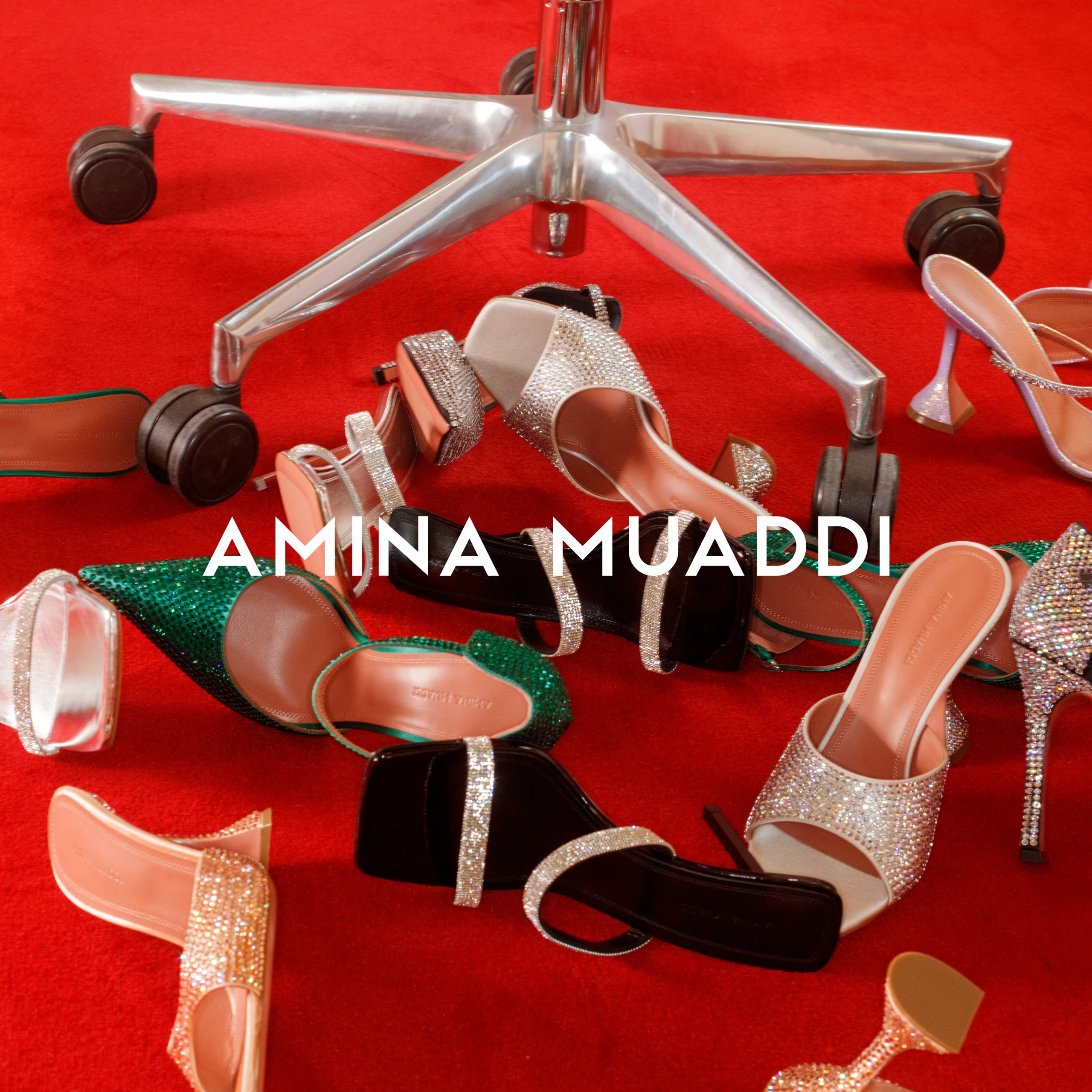 Collection of Amina Muaddi crystal-embellished heels scattered on red carpet around chair base