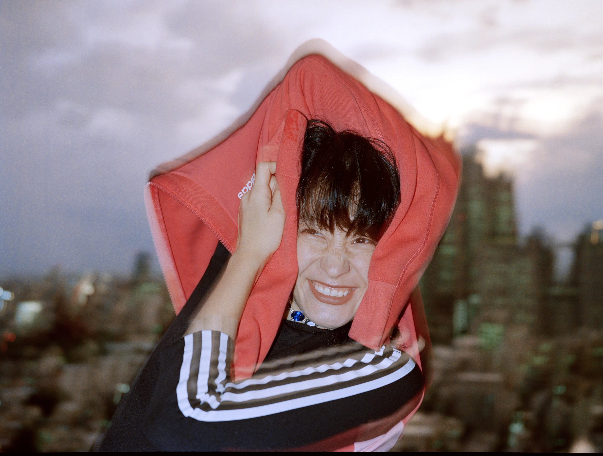 Athlete pulling red adidas hoodie over head smiling against Tokyo dusk skyline