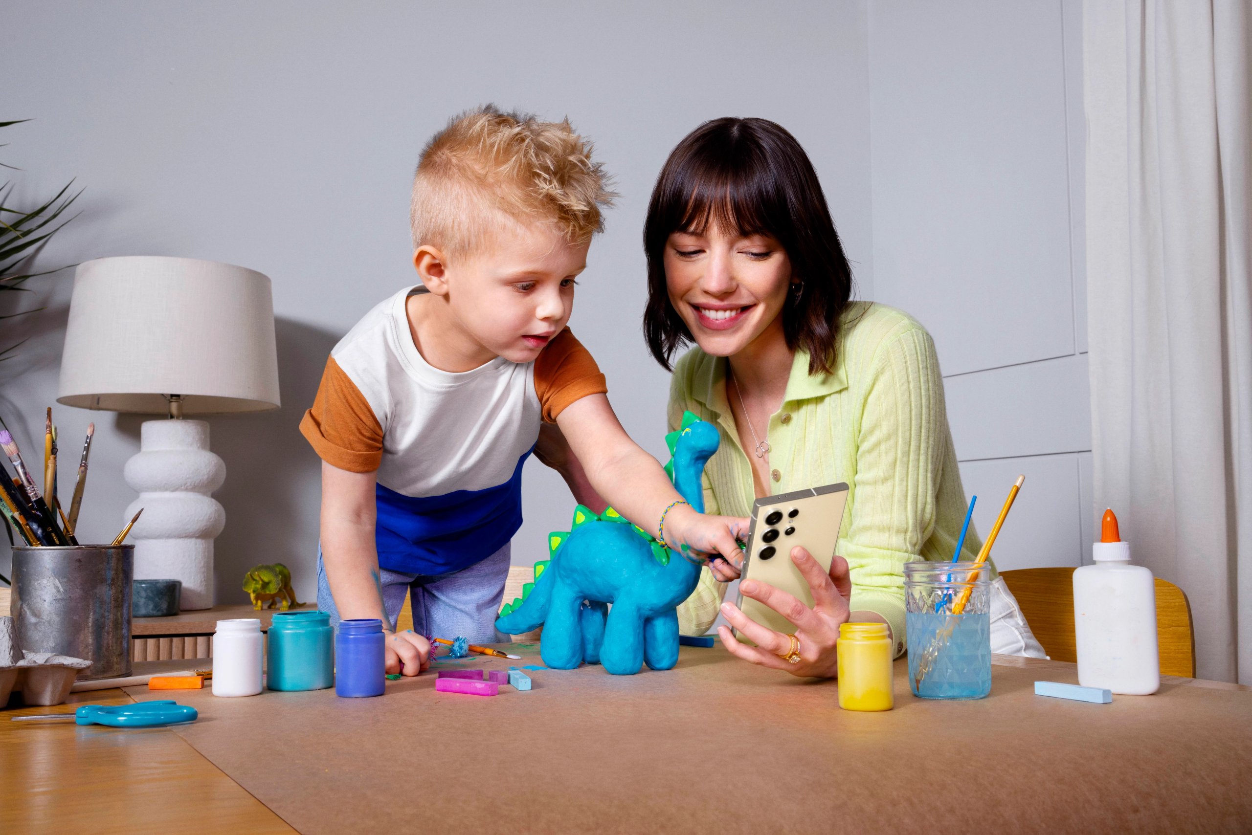 Mother and son crafting with Samsung Galaxy phone photographing toy dinosaur at craft table