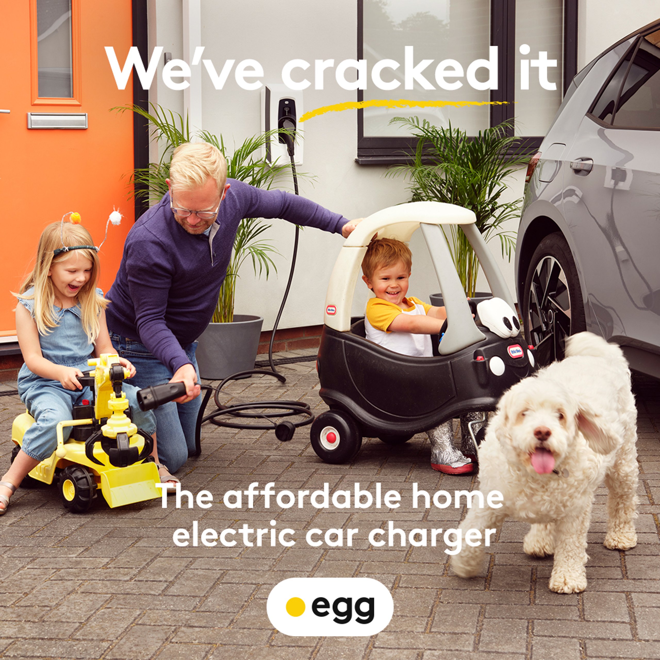 Father and children pretend-charging toy cars on driveway with EGG home charger and dog for Cracked It campaign