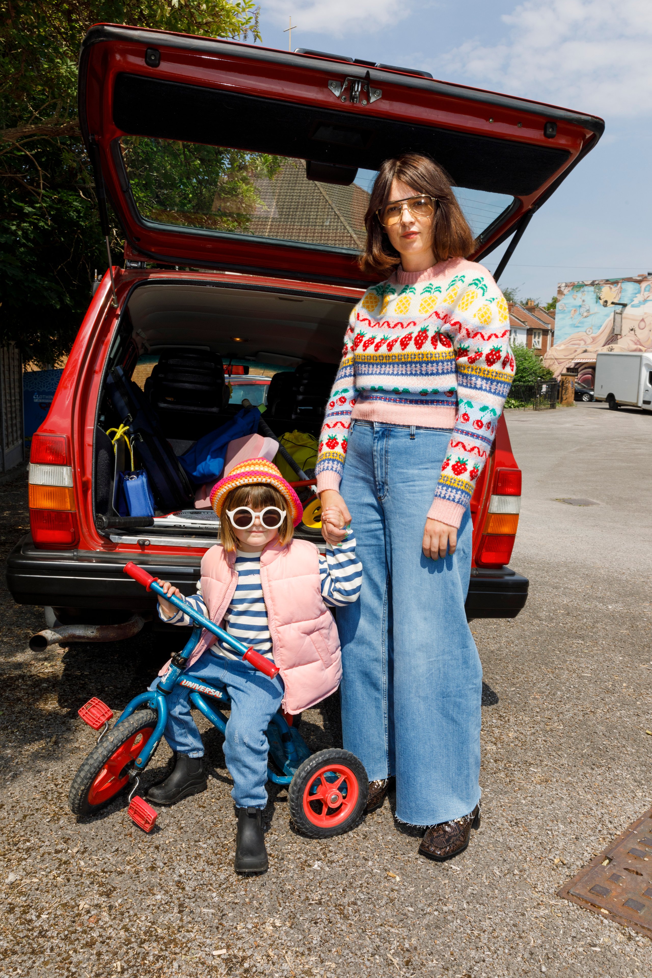 Mother and toddler with tricycle by red car boot in patterned knitwear and pink puffer jacket
