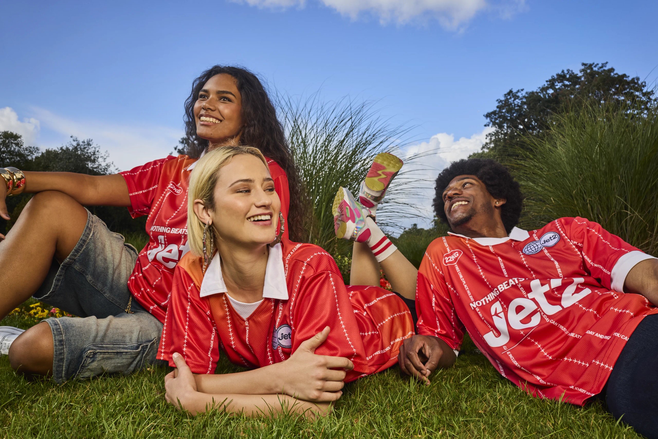 Three models in red Jet2 branded football jerseys lounging on grass in park