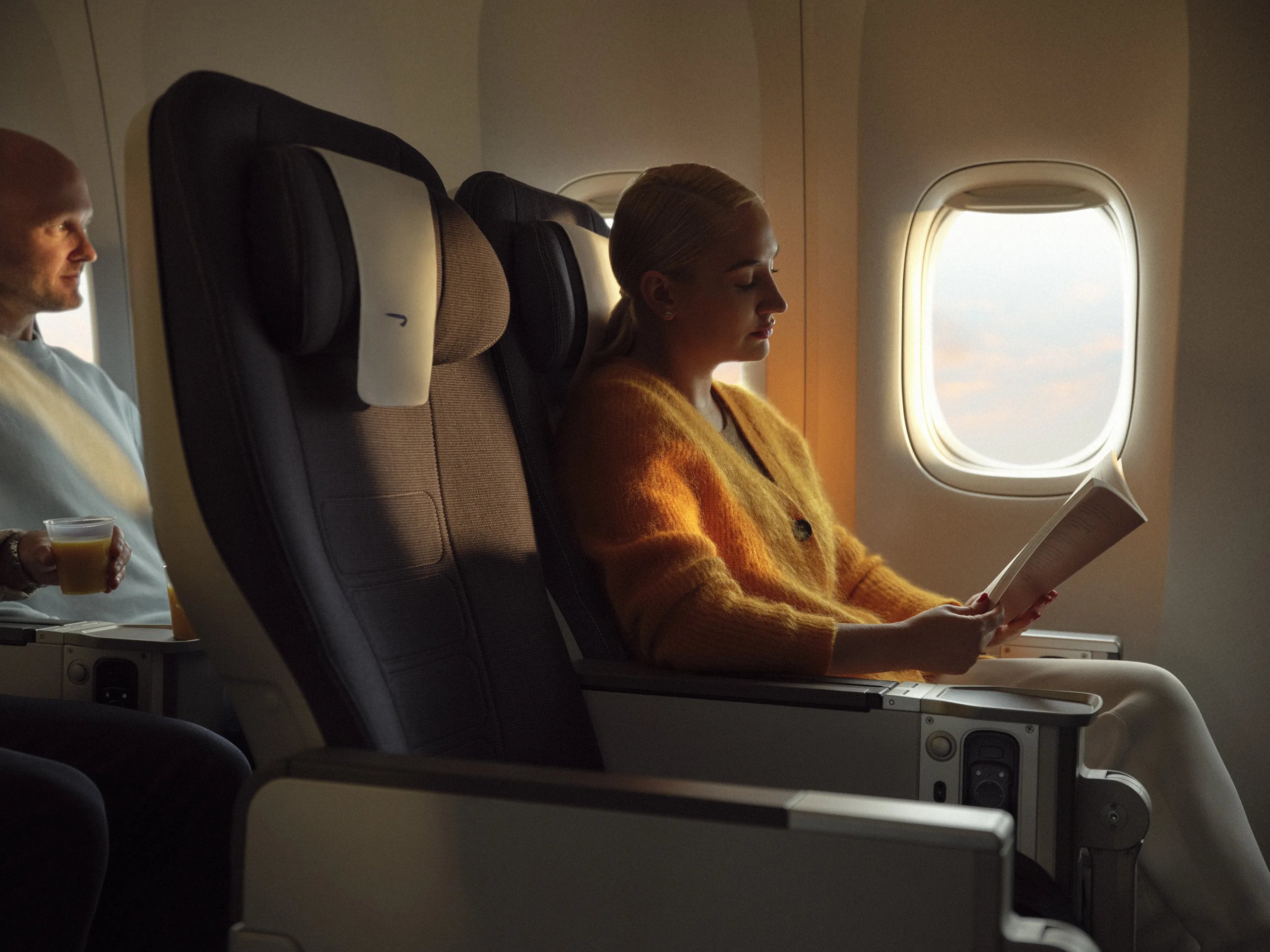 Woman reading book in British Airways premium economy seat in mustard cardigan with sunset window light