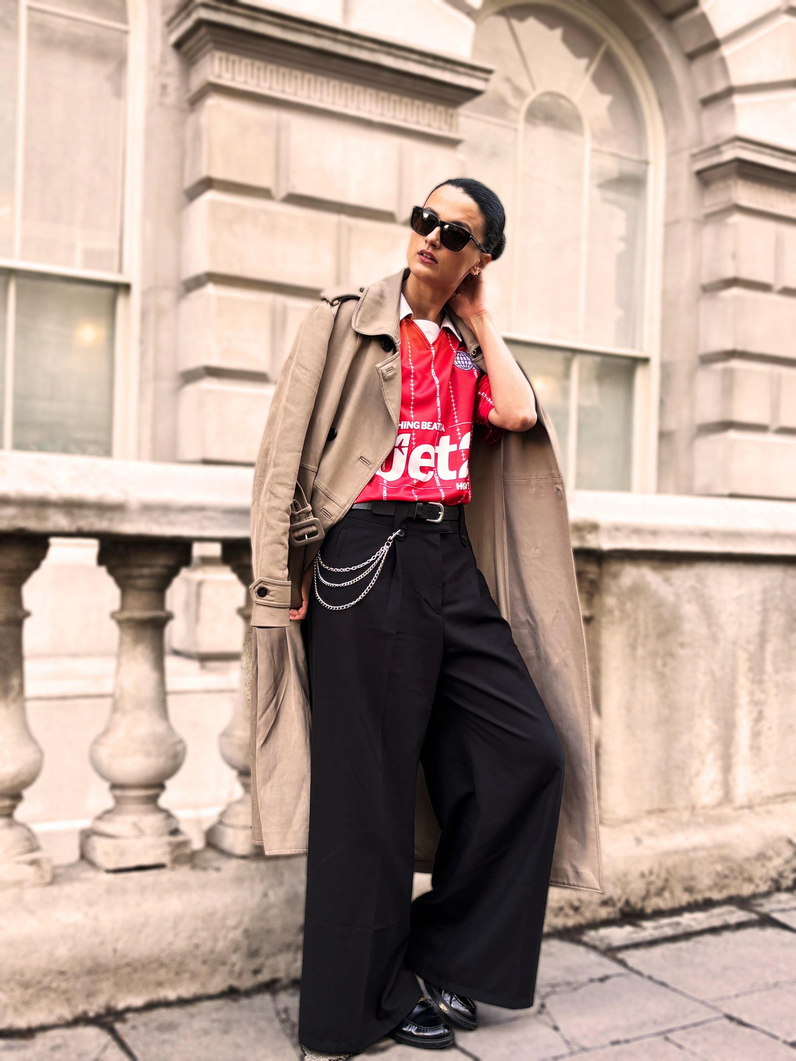 Model in beige trench coat over red Jet2 jersey with chain belt and wide trousers at Somerset House