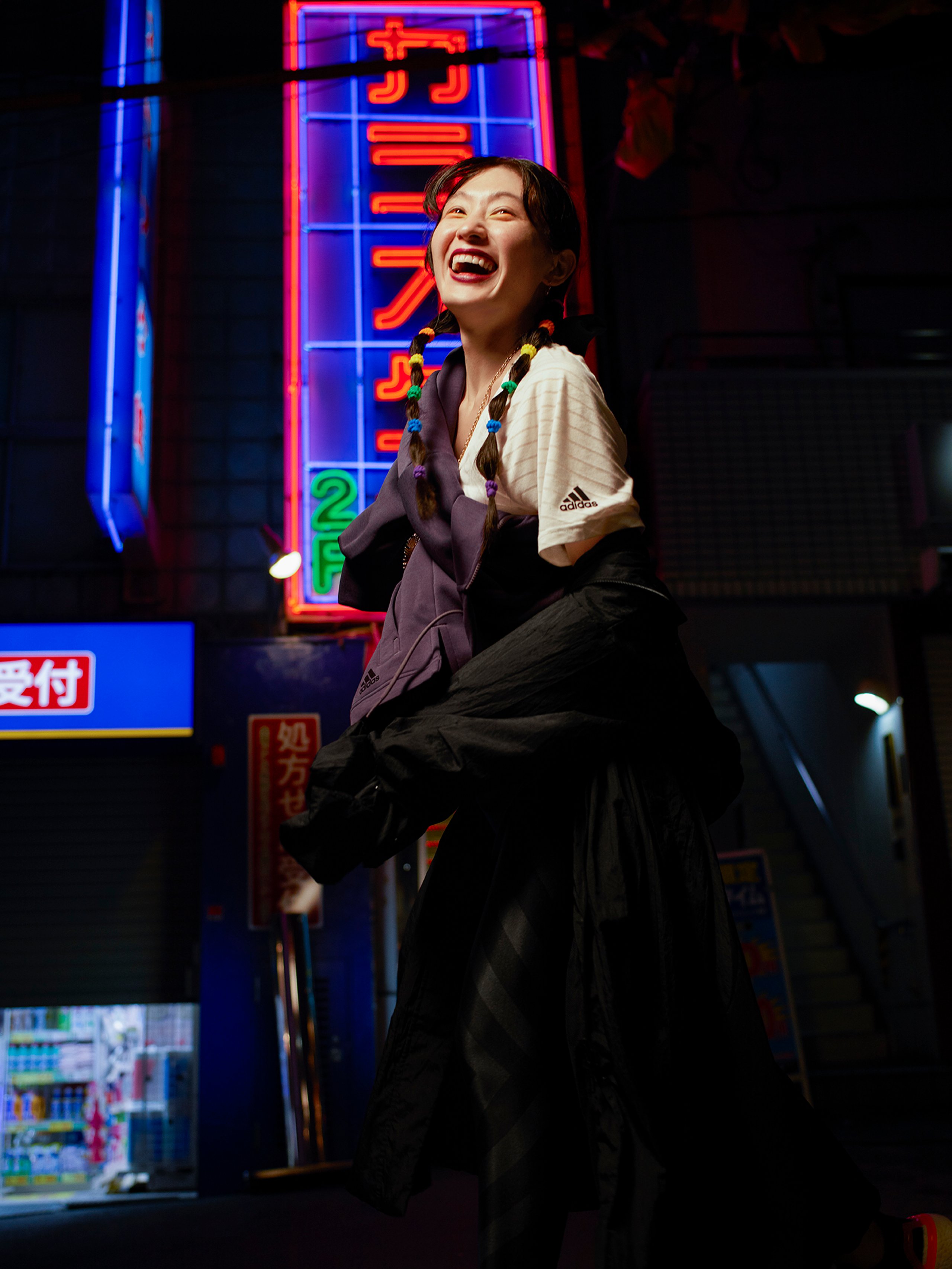 Athlete laughing in adidas tee and black skirt with colourful braids under Tokyo neon signage
