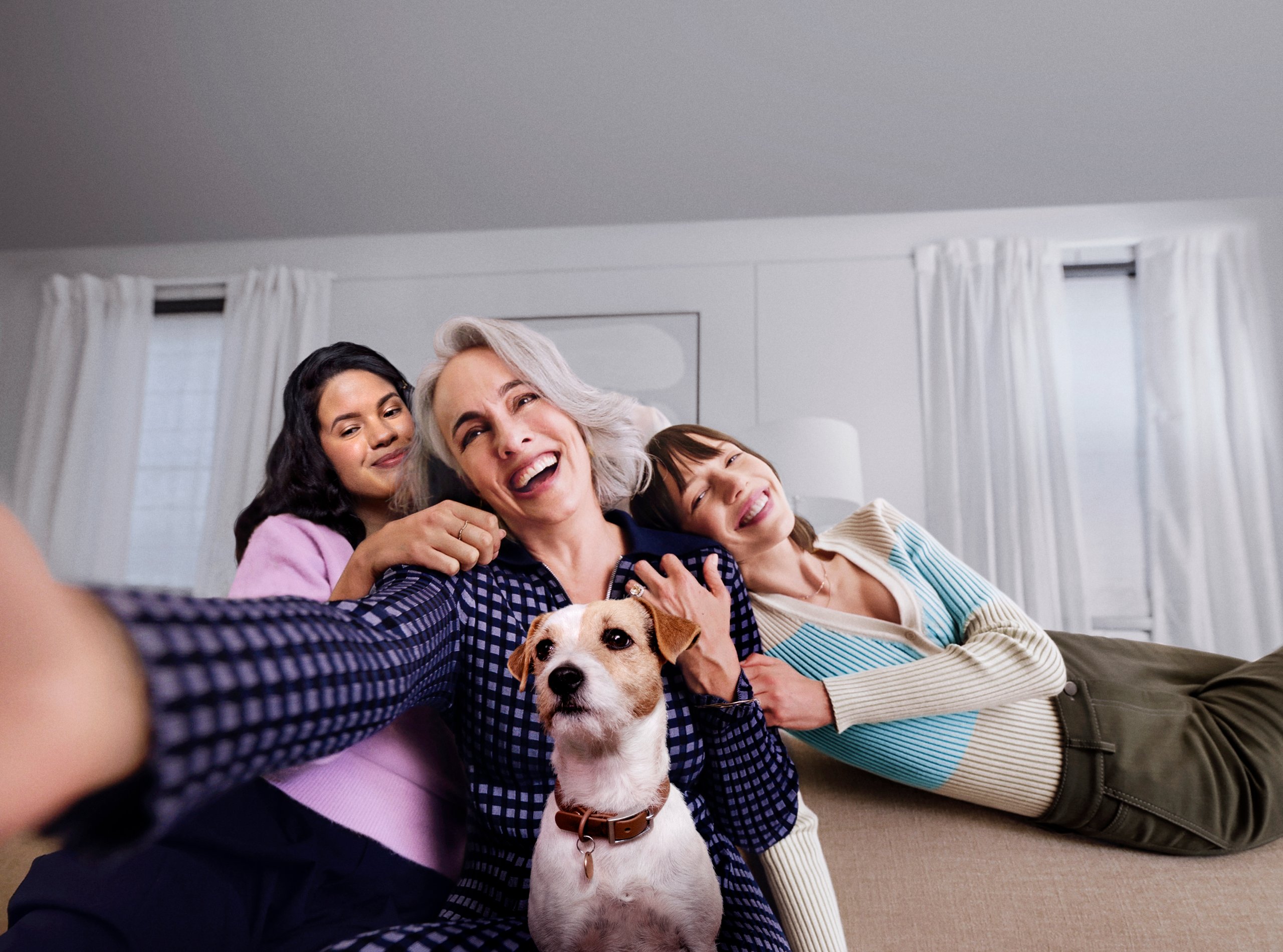 Three generations of women laughing together on sofa taking selfie with Jack Russell terrier