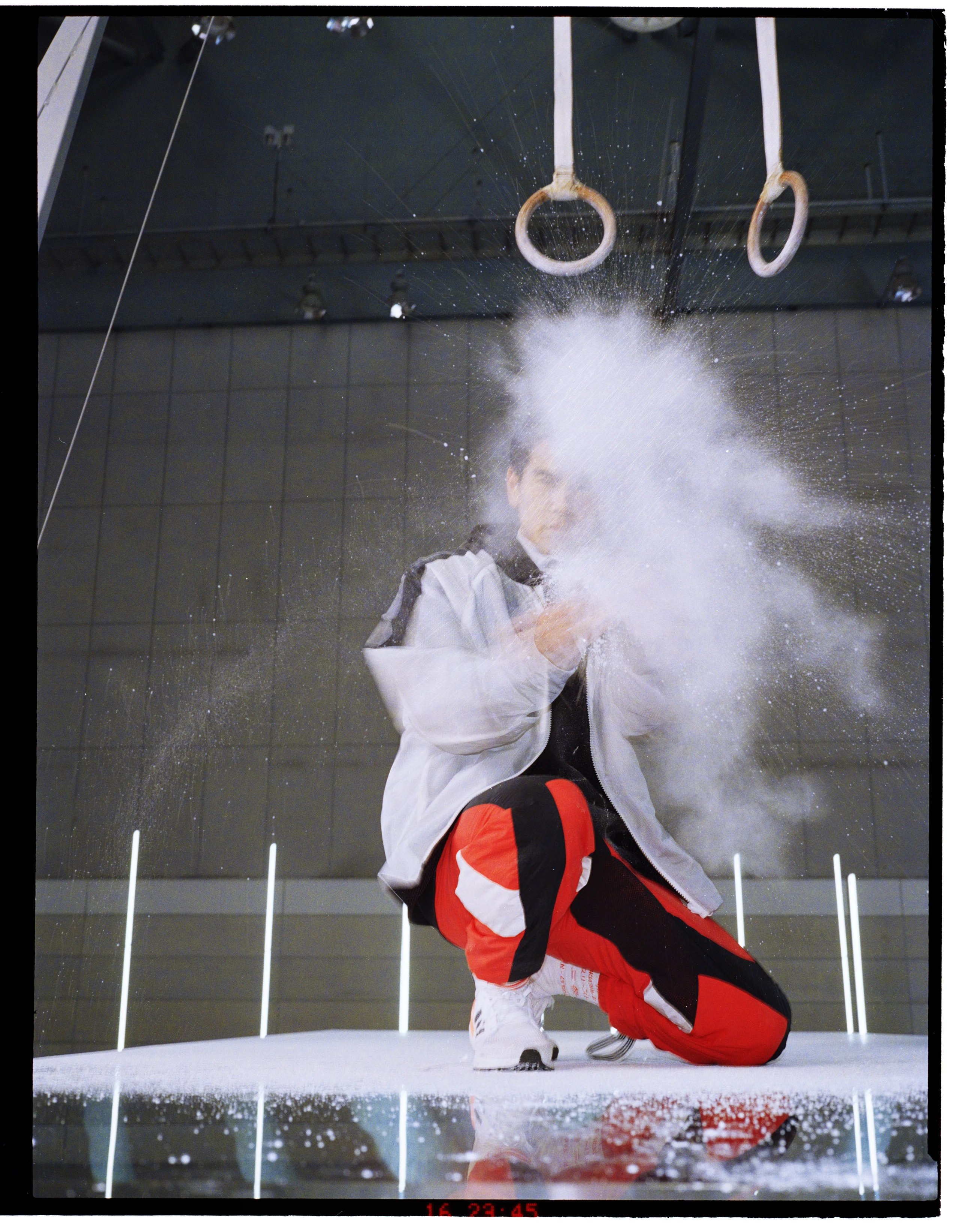 Gymnast clapping chalk dust in white adidas jacket and red leggings crouching under gymnasium rings