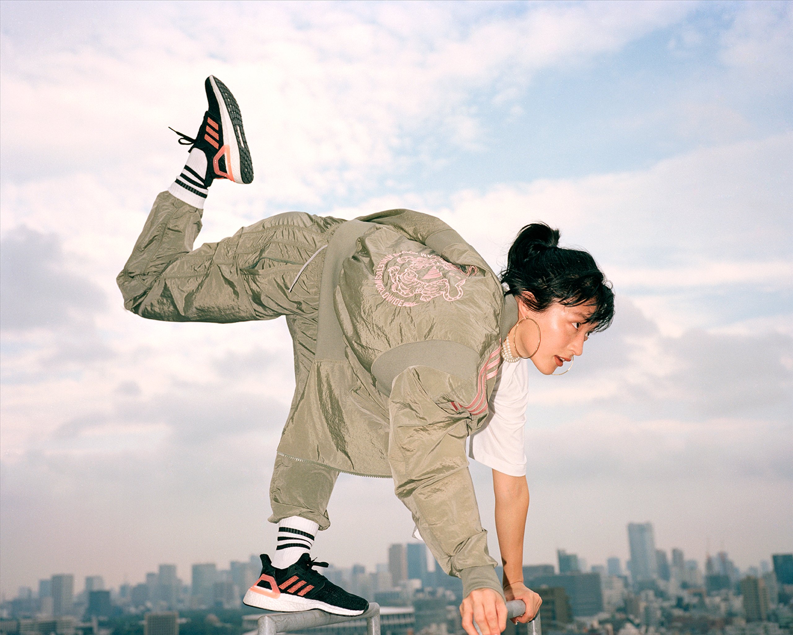 Athlete in olive bomber jacket doing handstand on rooftop railing with adidas Ultraboost trainers and Tokyo skyline