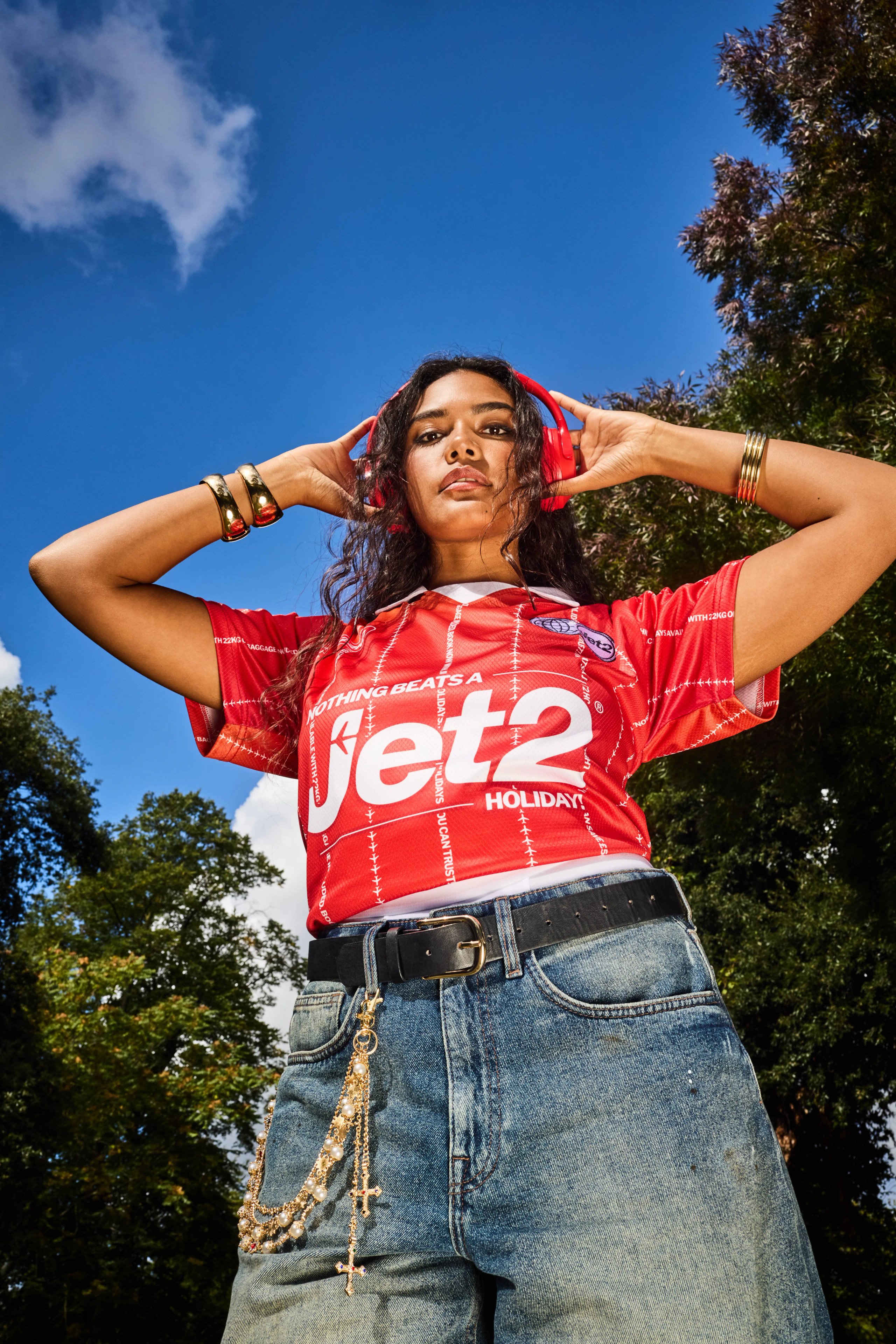 Model in cropped red Jet2 football jersey with red headphones, gold bracelets and denim jeans