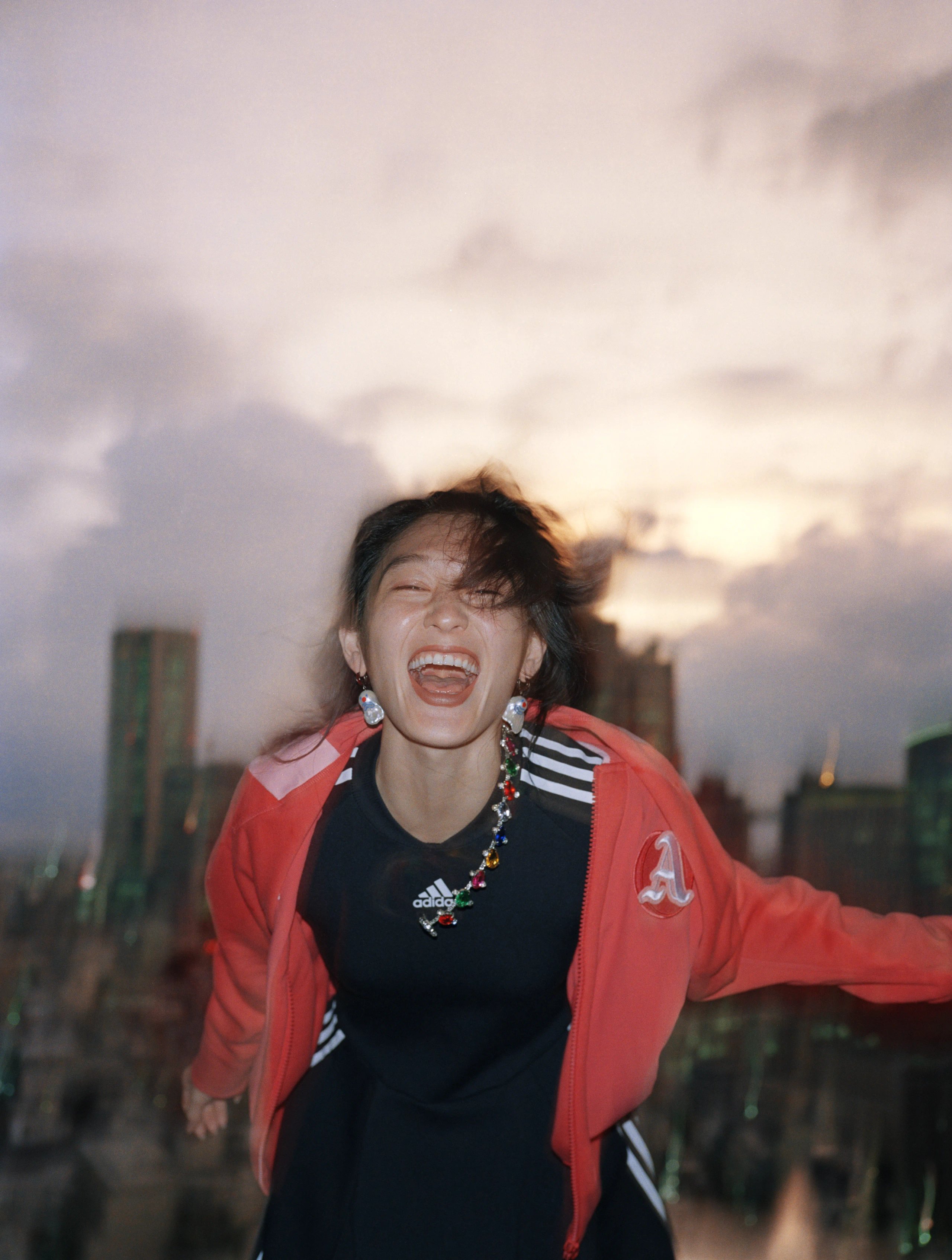 Athlete laughing in red adidas track jacket with jewelled necklace against Tokyo sunset skyline
