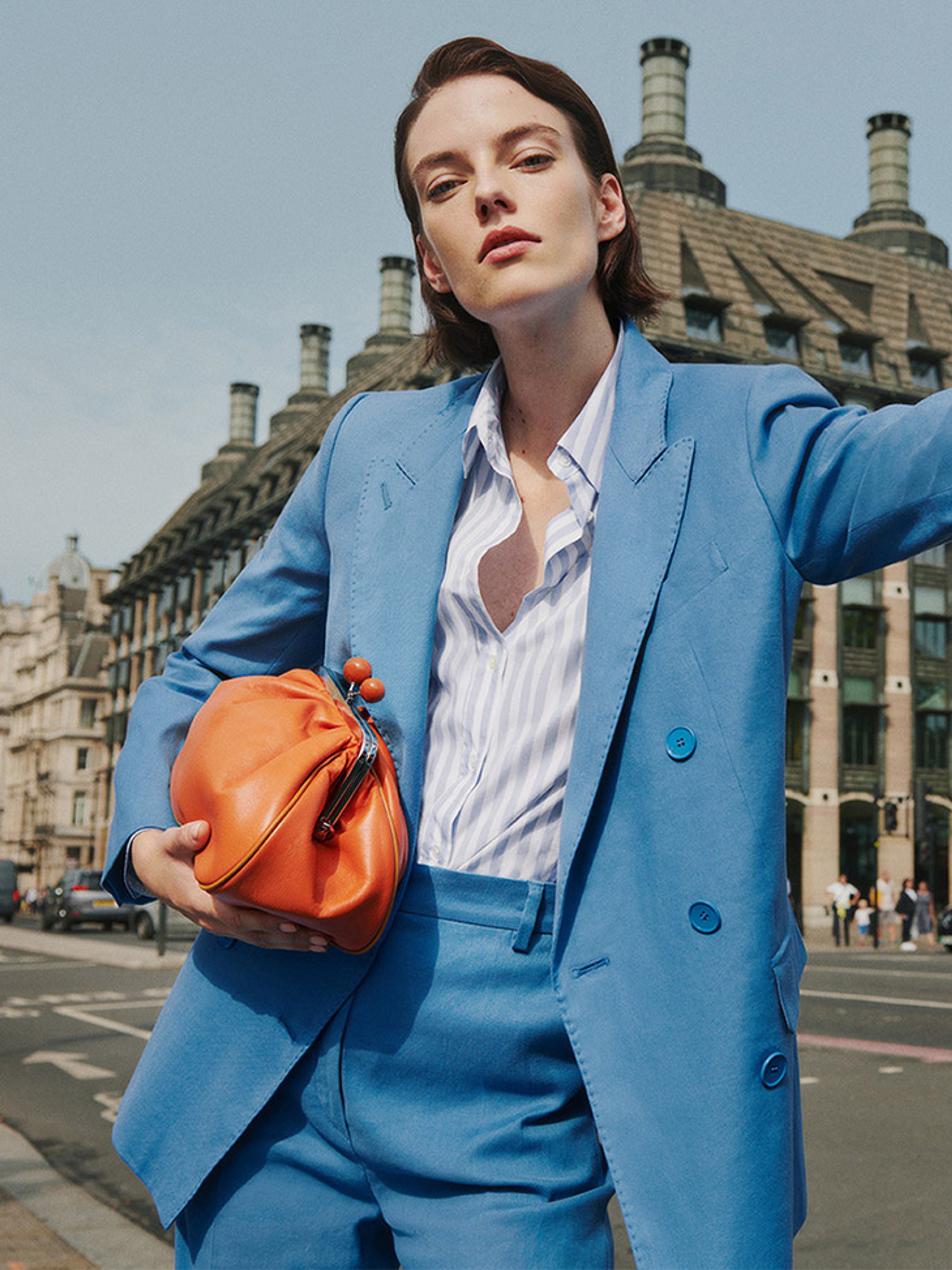 Model in sky blue double-breasted suit with striped shirt and orange leather clutch on London street