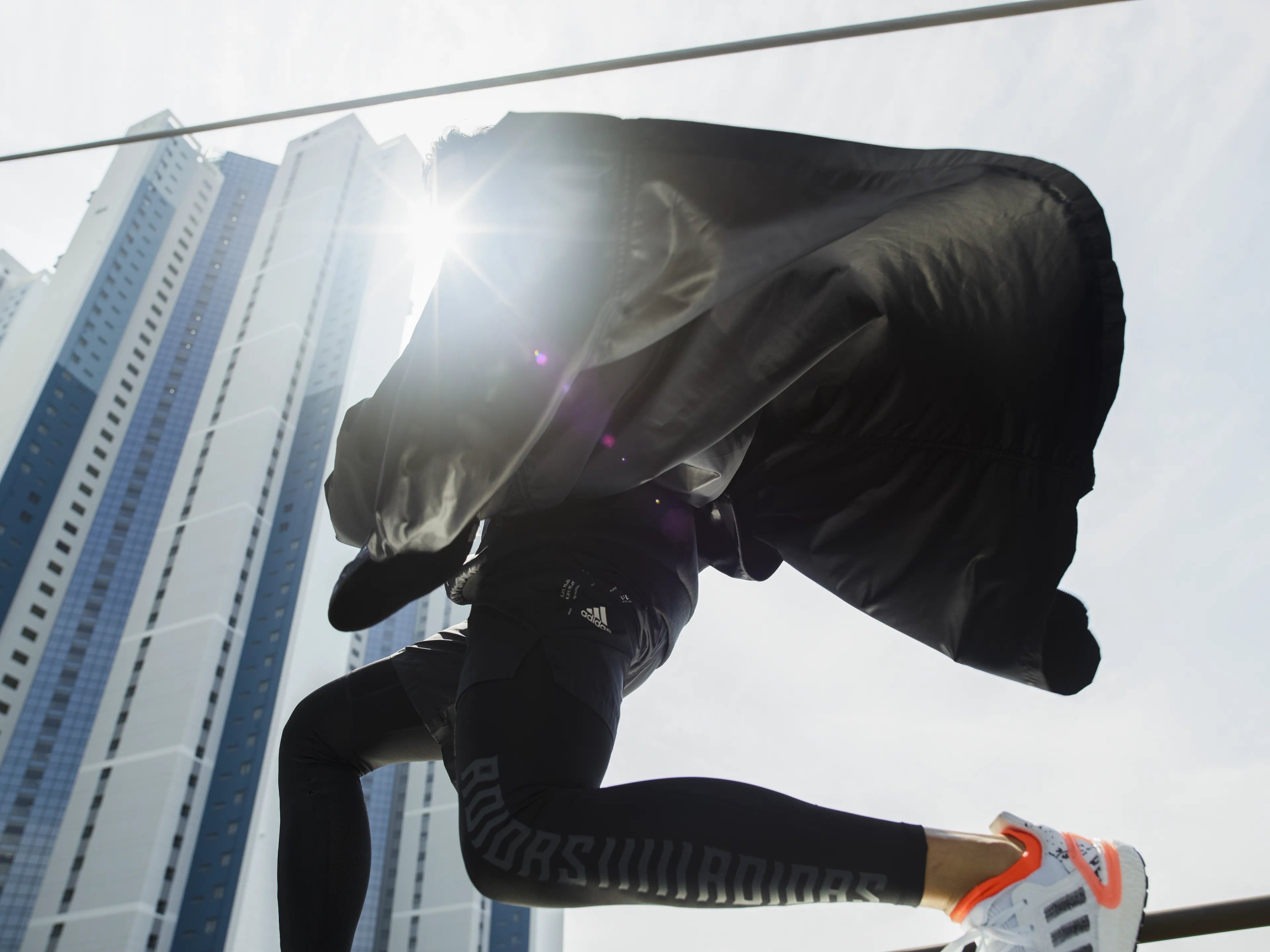 Athlete mid-stride silhouetted against sun flare with billowing adidas jacket and city skyscrapers