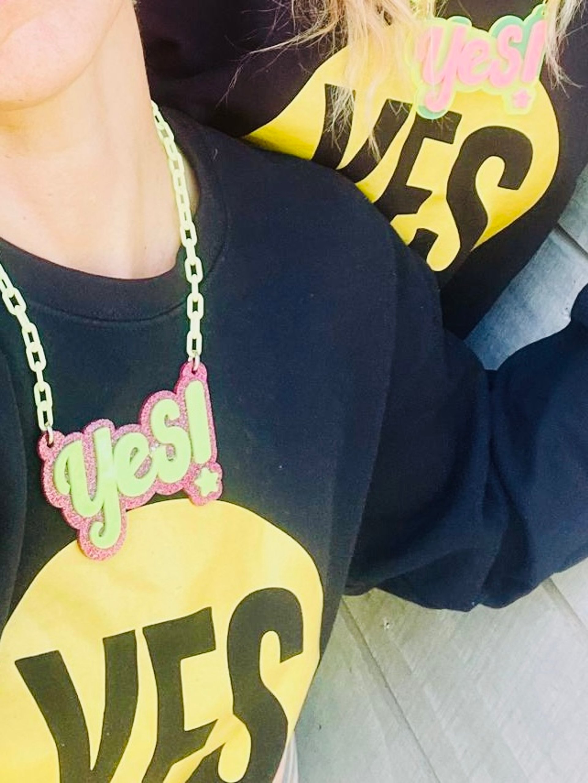 Close-up of YES! charity acrylic necklace in pink glitter and green over black hoodie with yellow YES! logo