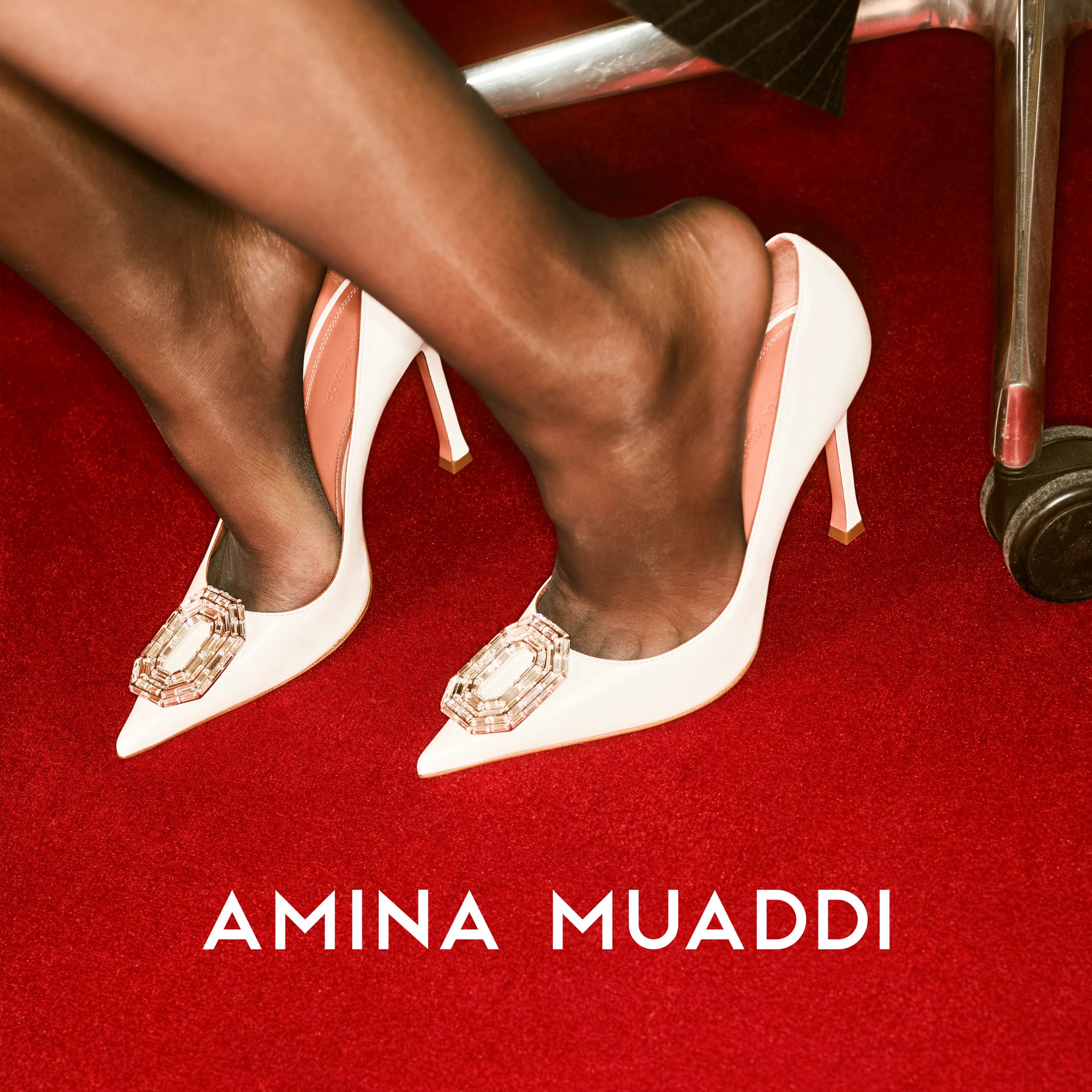 Amina Muaddi white satin pumps with crystal buckle on red carpet by office chair