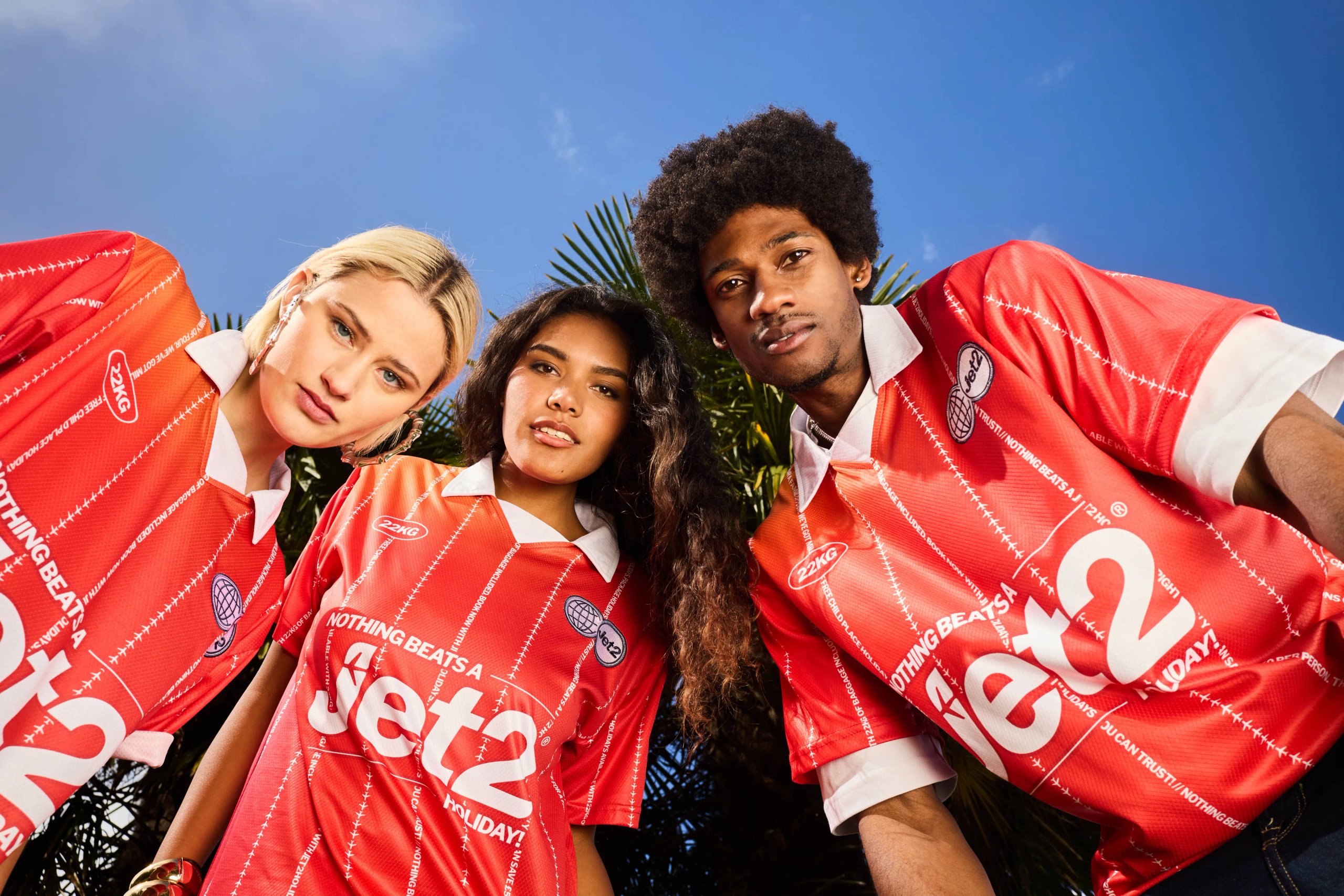 Three models in red Jet2 football jerseys shot from below against blue sky and palm trees