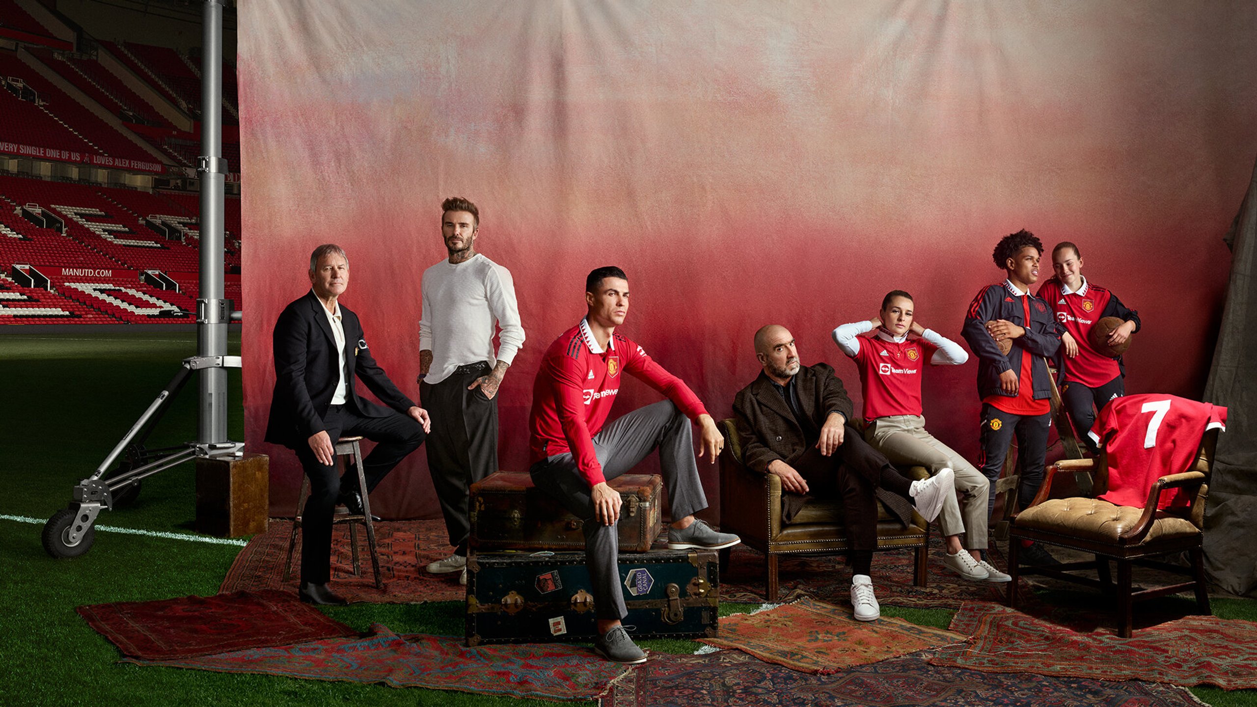 Manchester United legends group portrait at Old Trafford with red gradient backdrop and number 7 shirt