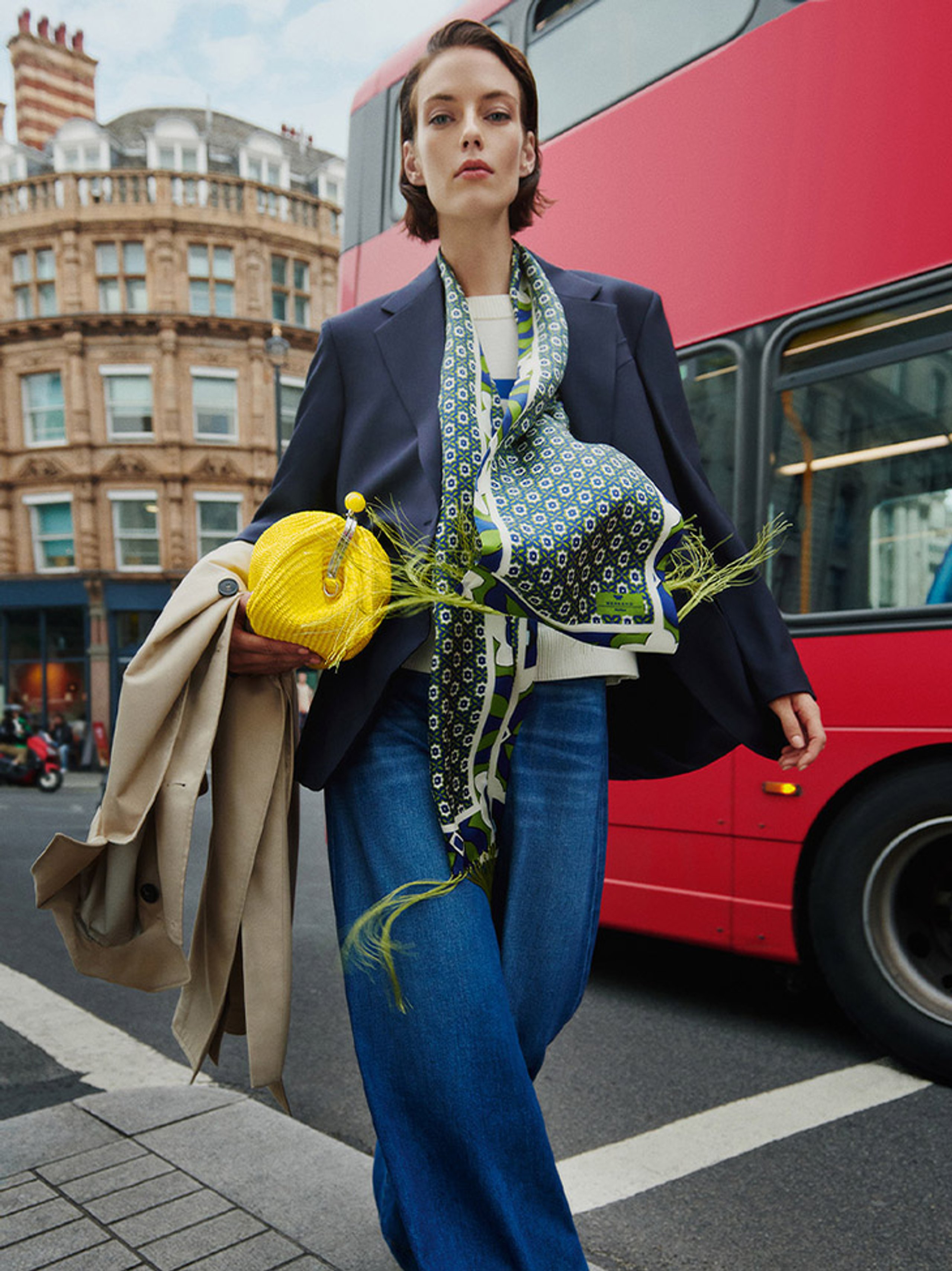Model in navy blazer with printed scarf and yellow clutch carrying camel trench coat by red London bus