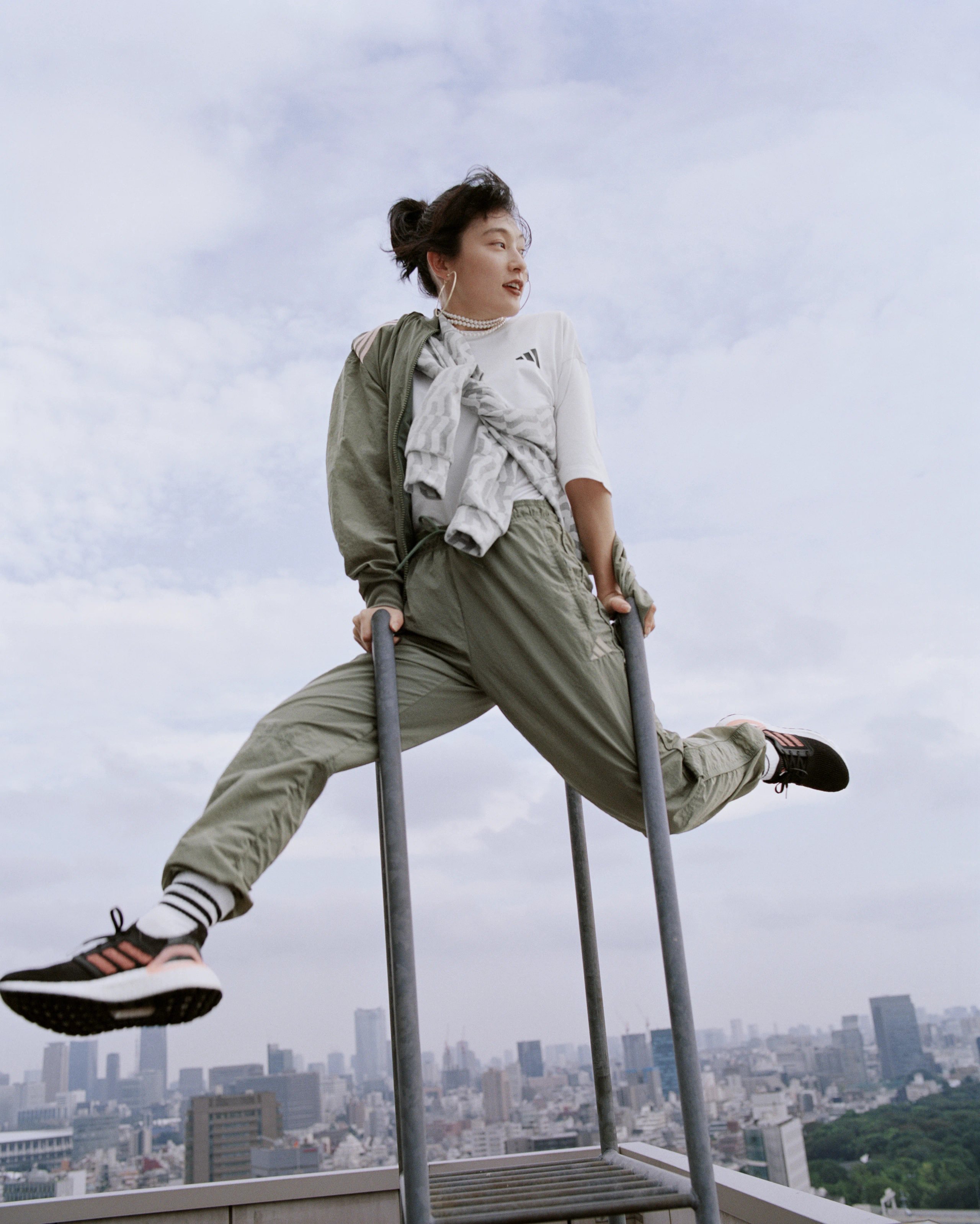 Athlete perched on rooftop railing in olive adidas tracksuit and Ultraboost trainers above Tokyo skyline