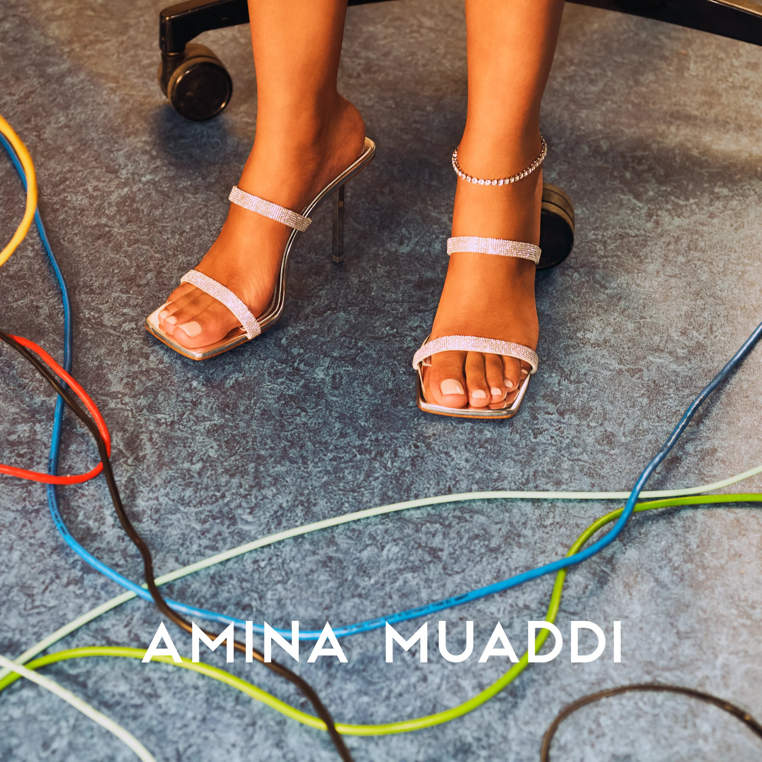 Amina Muaddi crystal strap sandals on grey floor surrounded by colourful cables