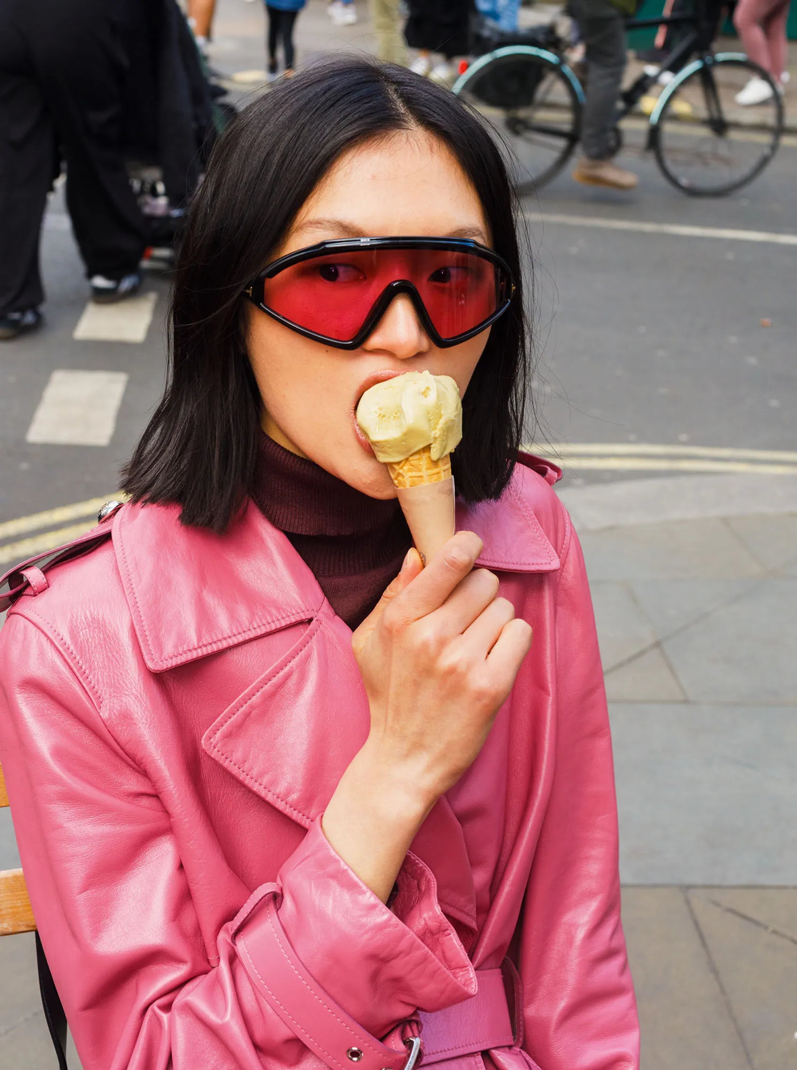 Model eating ice cream cone in pink leather trench coat and red visor sunglasses