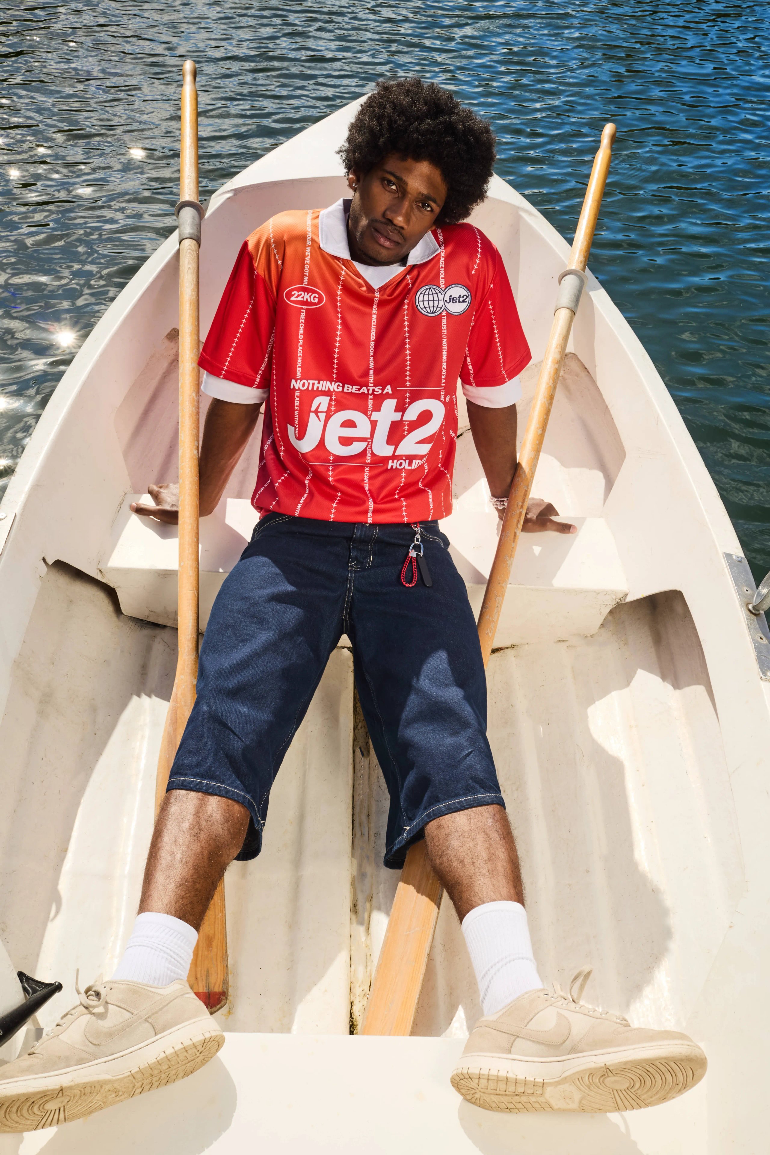 Male model in red Jet2 football jersey sitting in rowing boat with oars on lake