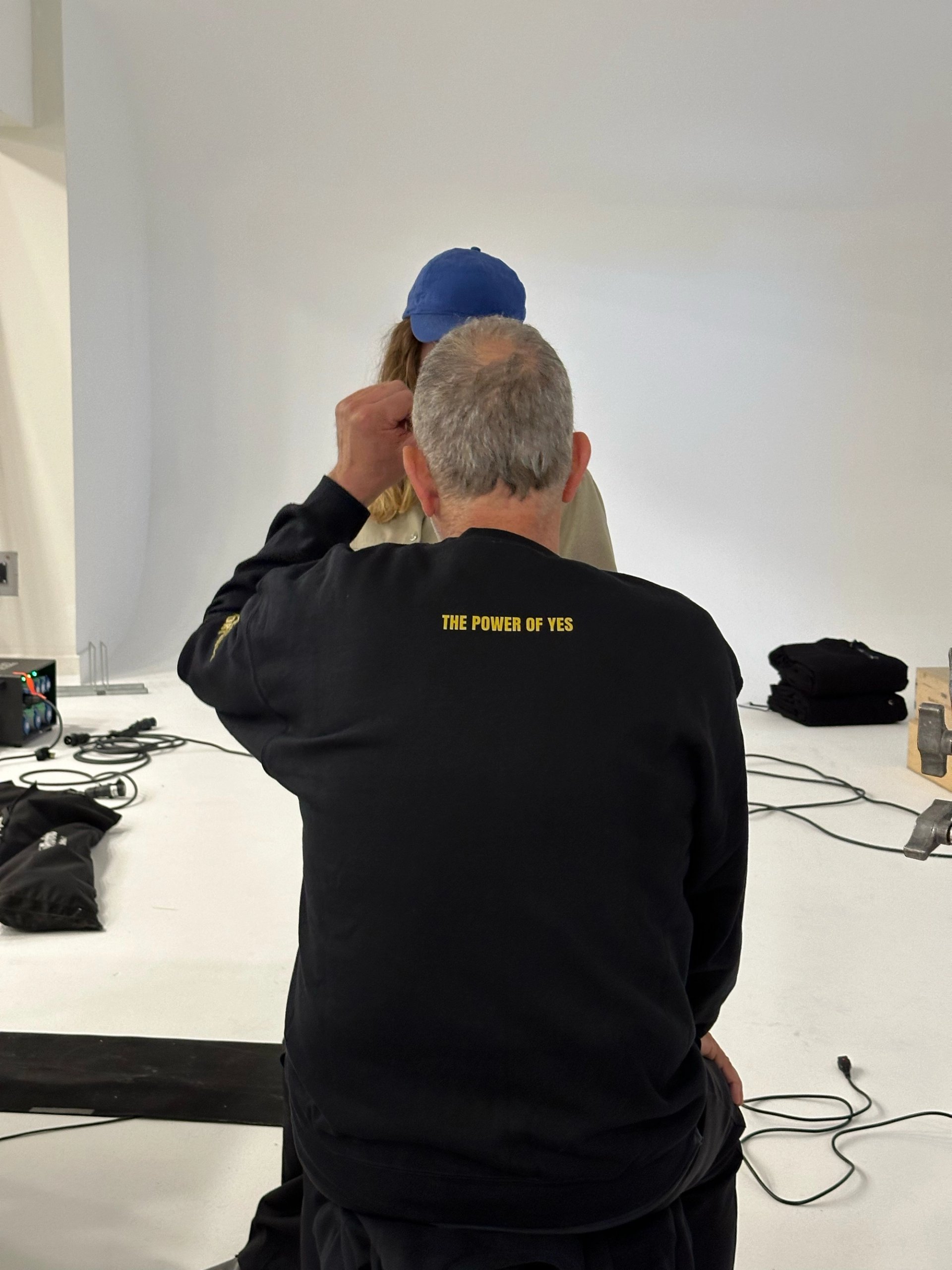 Photographer in The Power of Yes crew sweatshirt directing model in white studio from behind
