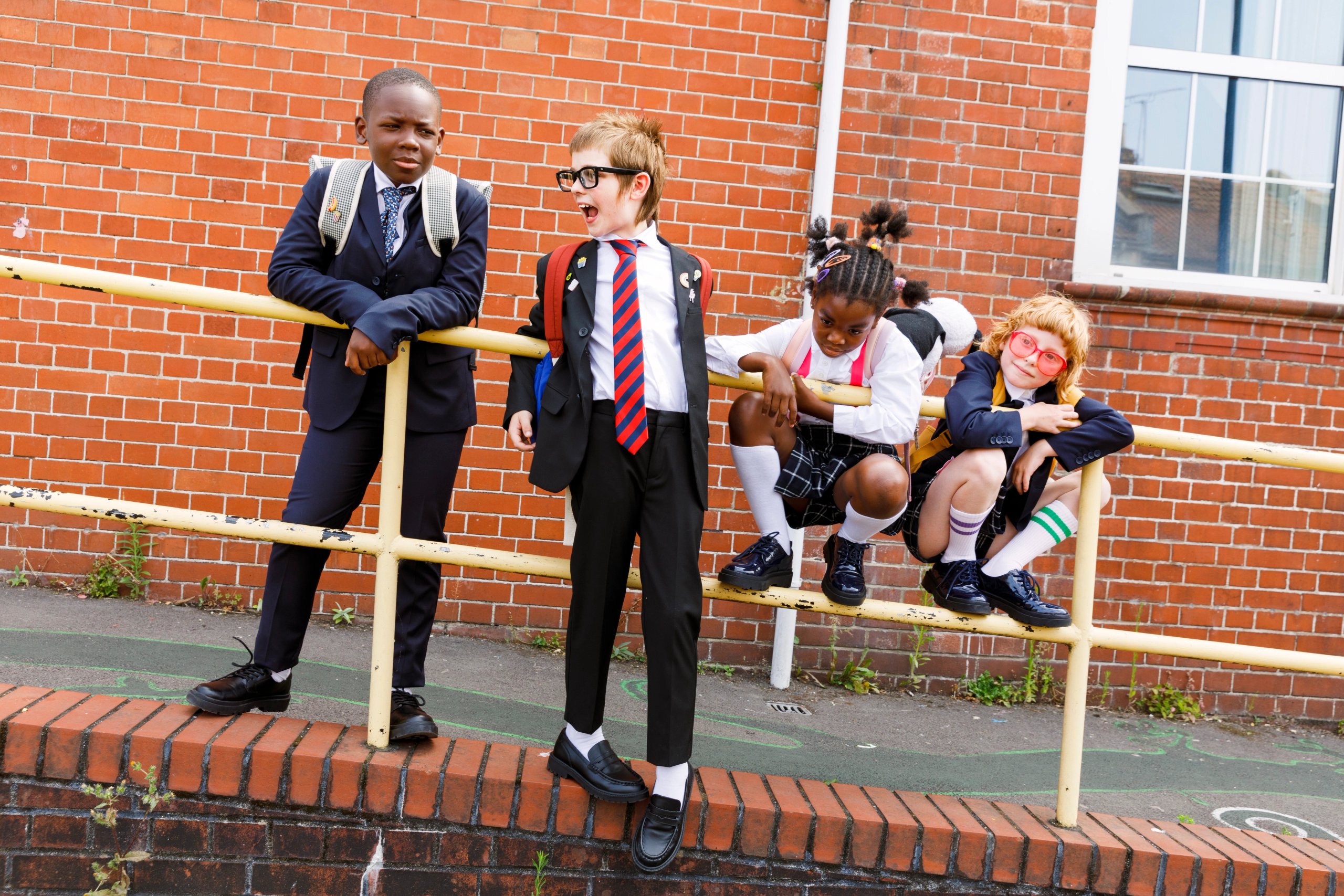 Four school children in navy uniforms laughing on yellow railing by brick wall